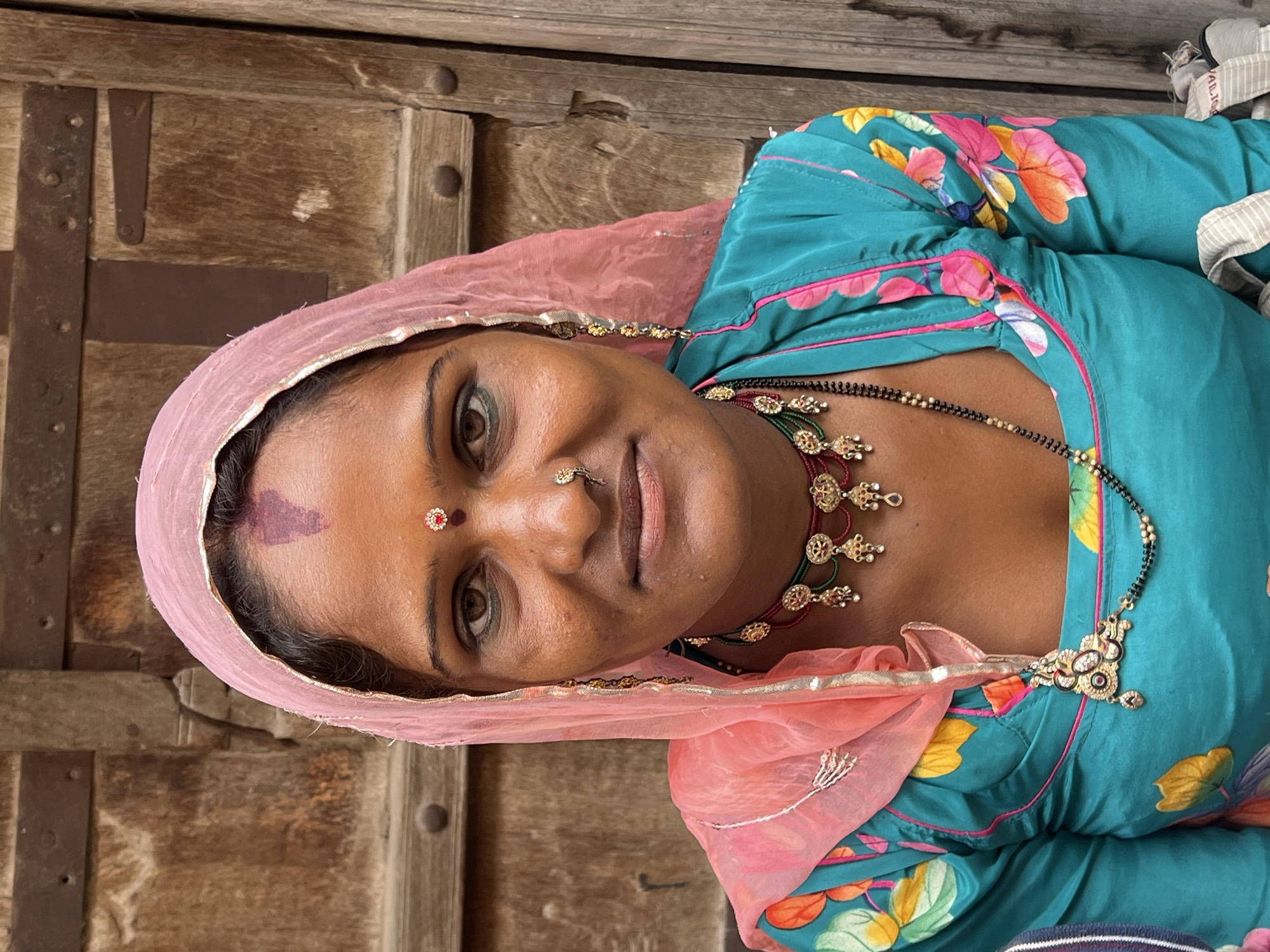 A woman with dark hair, brown eyes, and a light pink headscarf is photographed from the chest up. She wears a turquoise blouse with colorful floral patterns on the sleeves. She is adorned with several pieces of jewelry, including a multi-strand beaded necklace, a choker with dangling pendants, earrings, and a nose ring. A red bindi is visible on her forehead, and a smaller red gem is placed above it. The background consists of a weathered wooden door. The image appears to have been taken outdoors in Pushkar, India, given the style of dress and jewelry.