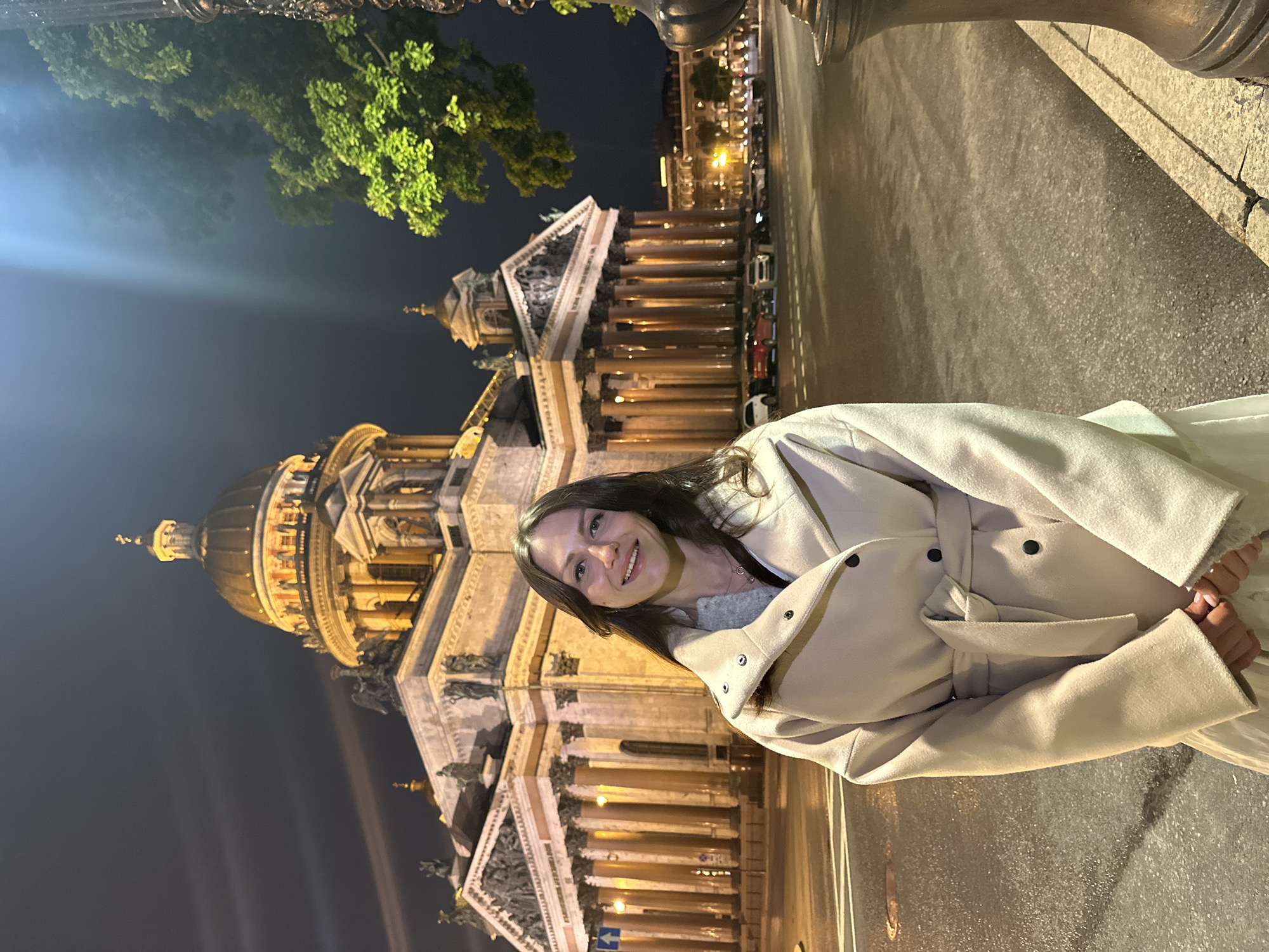 A woman in a light-colored coat stands in front of Saint Isaac's Cathedral in Saint Petersburg, Russia at night. The cathedral is illuminated, and there is a tree with green leaves visible in the upper left corner. The pavement is visible on the right side of the image.