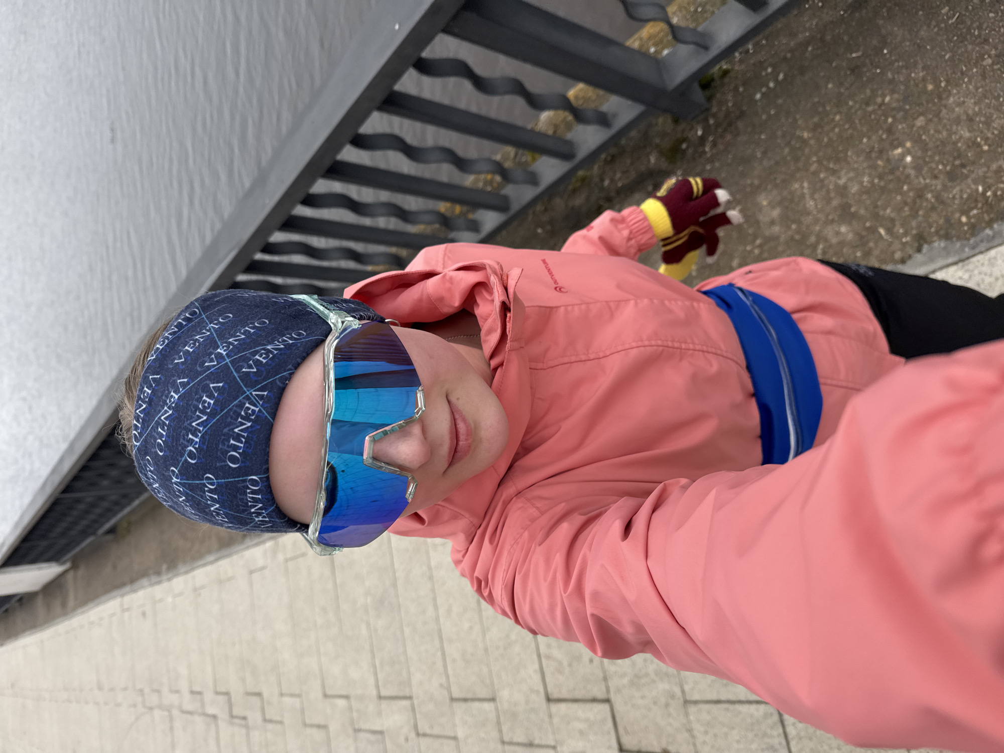 This is a tilted selfie featuring a person outdoors, likely in Chaikovsky, Russia, during the daytime. The individual, seen from a slightly elevated perspective, holds the camera at arm's length. They are dressed in active wear, including a salmon-pink jacket, a bright blue fanny pack or belt bag worn across the waist, and a dark blue headband patterned with the word "VENTO" repeated several times. Their eyes are obscured by large, reflective blue sports sunglasses. On their right hand, they wear a maroon and yellow striped glove with white fingertips.
The background shows a light grey, textured building wall on the left. To the right and behind the person, a dark grey metal railing or structure with wavy bars is visible, with some darker, rough ground beneath it. The immediate ground surface appears to be paved with light-colored concrete or flagstones. The lighting is bright and even, suggesting an overcast day, and there are no strong shadows. The primary activity is taking a selfie during an outdoor outing, possibly for recreation or sport, given the attire.
Visible text includes "VENTO" on the headband and "OUTVENTURE" on the right side of the pink jacket.