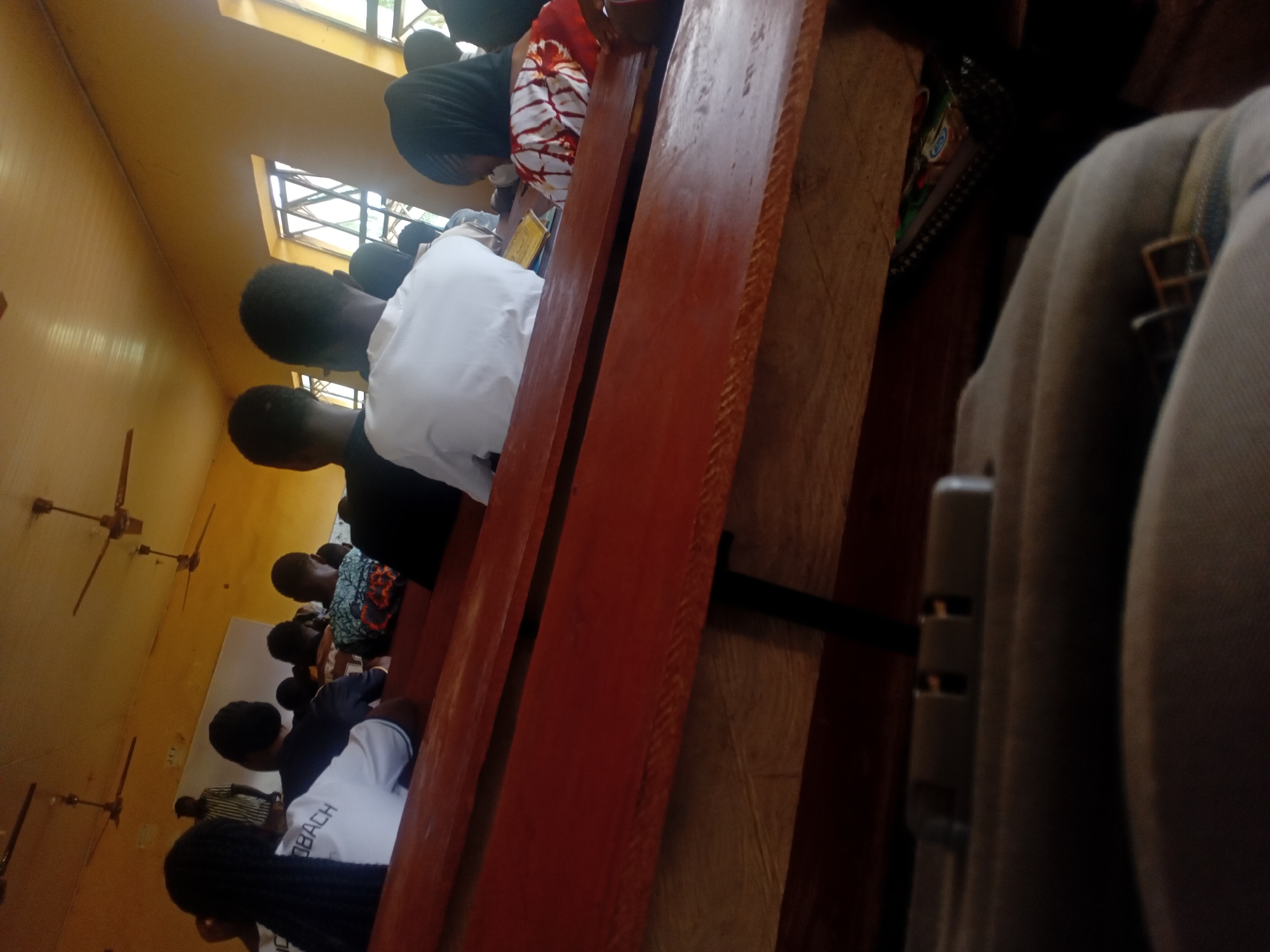 A partially obscured indoor scene captures a row of people seated in what appears to be a church or similar religious setting. The image is taken from a vantage point that's skewed, with the view oriented diagonally across the rows of seated individuals. A prominent wooden bench or pew stretches across the middle ground, with the back of the bench rising higher in the frame.
The individuals are of African descent, and the clothing they wear varies, from white shirts to patterned fabrics, suggesting different roles or attire requirements.
The environment shows a window allowing natural light to stream in, and ceiling fans are visible, indicating a need for ventilation in the space.
The location is labeled as Unknown, Unknown, providing no specific geographic information about the city or area where the photo was taken.