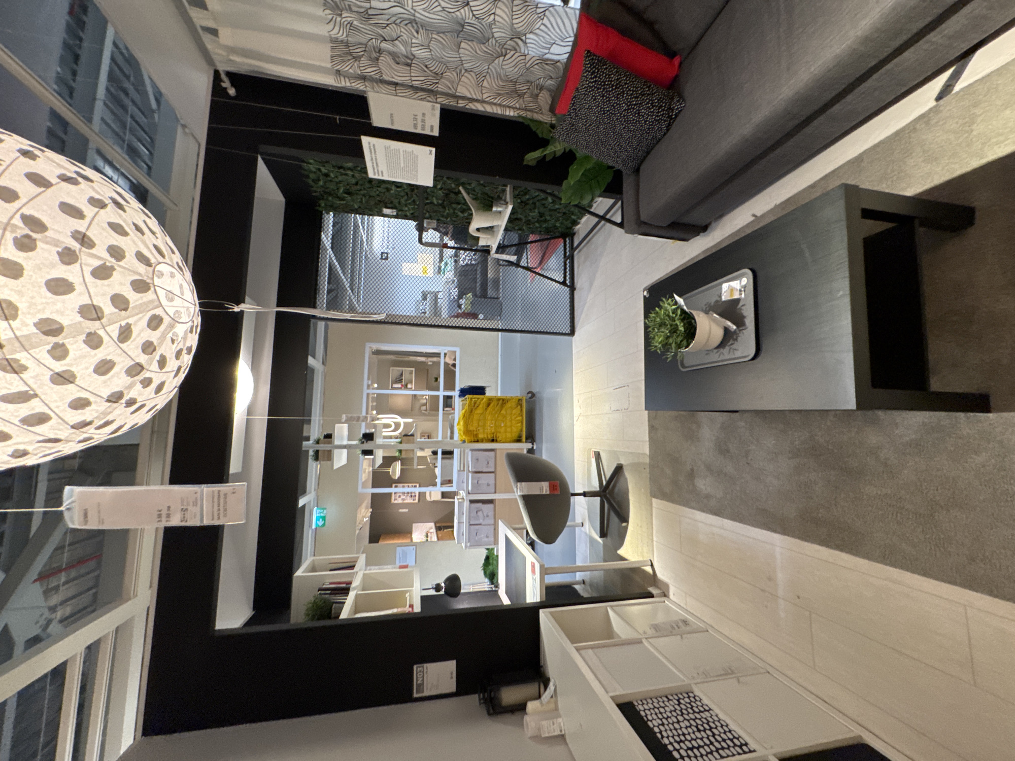 The image depicts an indoor scene, likely a showroom or retail space, with furniture and home decor items. The overall aesthetic suggests a modern and minimalist design. In the foreground, a dark gray sofa with two throw pillows, one red and one patterned black and white, is visible. A dark rectangular coffee table sits in front of the sofa, with a small potted plant and a tray on it.
To the left of the sofa, a light-colored wooden floor transitions to a gray rug. Further left, a white modular shelving unit is partially visible, filled with various items. Adjacent to the shelving unit is a black desk with a chair. The chair is a swivel chair with a gray upholstered seat and a dark base. A black desk lamp is positioned on the desk.
In the background, there is a display of white storage boxes and a bright yellow trolley. Further back, a doorway leads to another section of the space, where more shelving units and lighting fixtures are visible. The ceiling features exposed beams and lighting. A large, spherical light fixture with a patterned shade hangs from the ceiling, partially obscuring the upper part of the image. There are also several signs and price tags visible throughout the scene, typical of a retail environment. The presence of greenery, both in the potted plant and a vertical arrangement on a wall in the background, adds a touch of nature to the space. The overall lighting is bright and even, illuminating the displayed items. There are no people visible in the image. The location is identified as IKEA Sofia, Bulgaria.