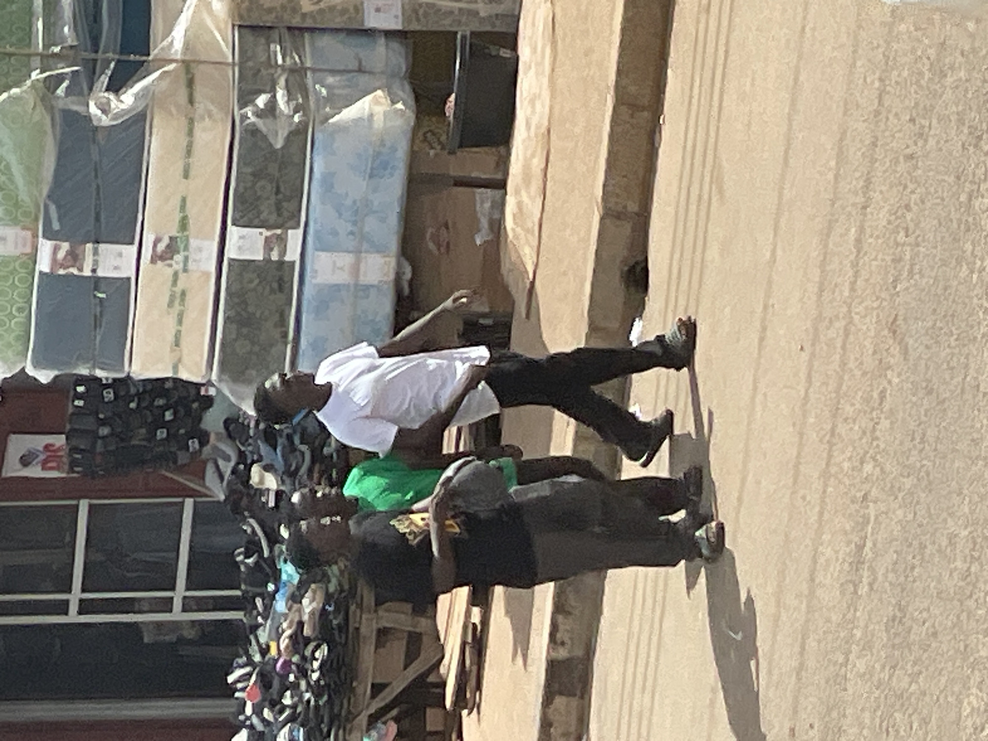 Three young men walk along a dusty street in front of a market stall. The man on the left wears a black t-shirt and grey pants, holding a dark object in his hand. The man in the middle wears a bright green t-shirt and appears to be walking. The man on the right wears a white t-shirt and black pants. Behind them, the market stall displays stacks of mattresses and various footwear. The environment is outdoor, likely in a tropical or subtropical climate, as suggested by the bright sunlight and dusty ground.