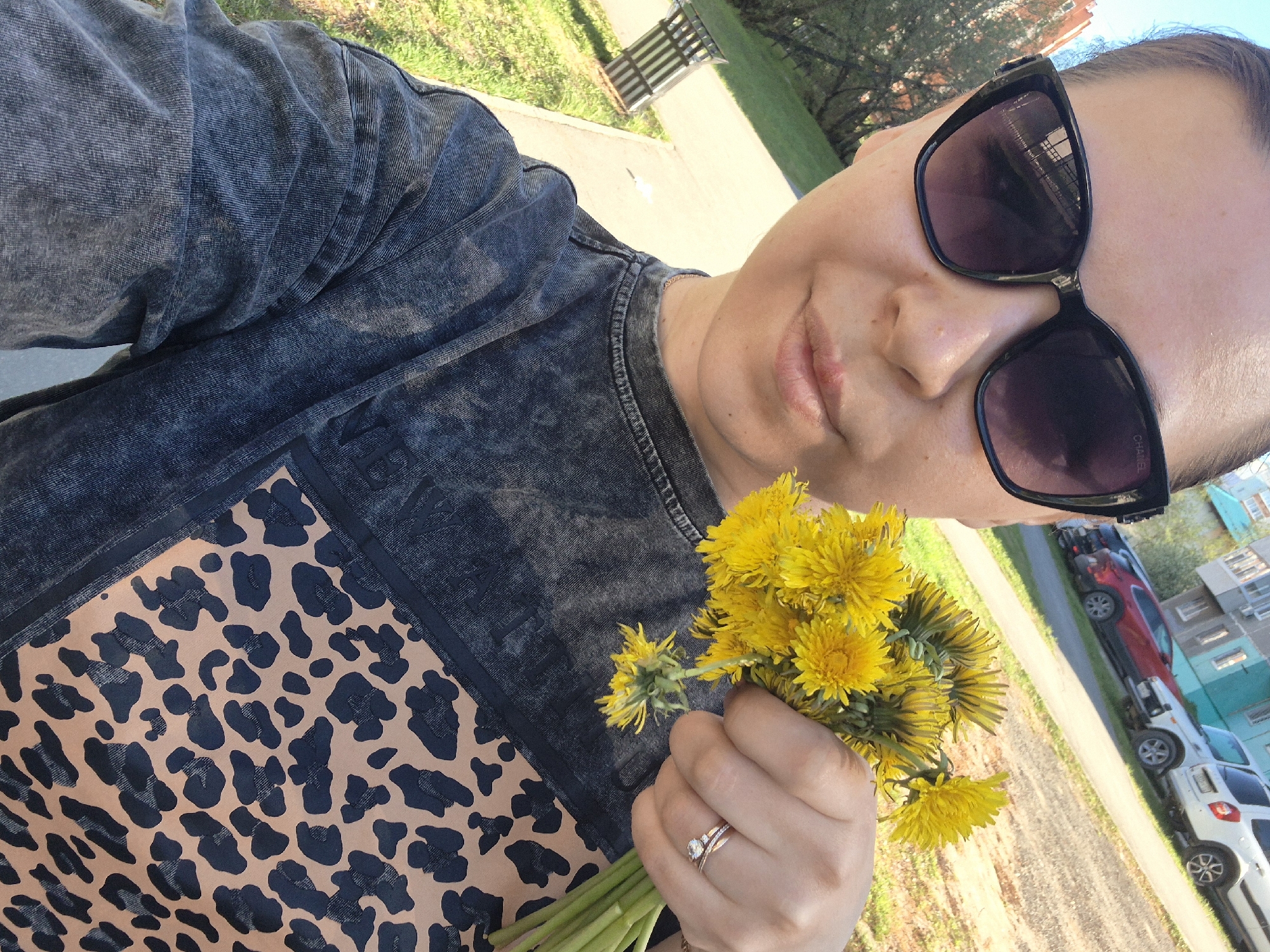 A woman wearing black-framed sunglasses and a gray shirt with a leopard print design is holding a bouquet of dandelions. She is wearing two rings on her left ring finger. Behind her, there's a sidewalk, grass, and a street lined with cars. Buildings are also visible in the background. The location is Tobolsk, Russia.