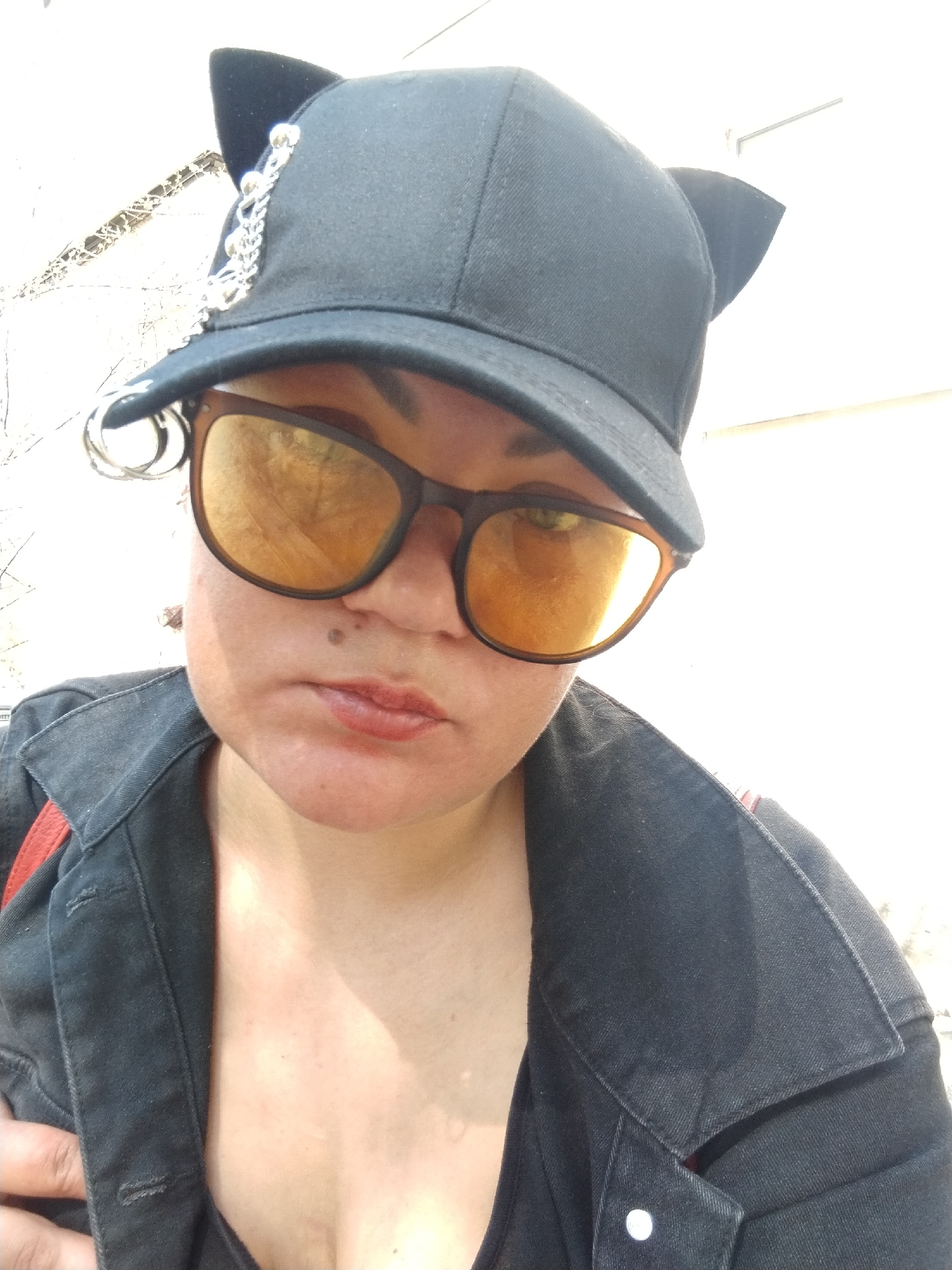 A close-up selfie shows a person wearing a black cap with cat ears, featuring a silver chain and rings on the brim, and amber-tinted sunglasses. They are also wearing a black denim jacket with a red strap visible underneath. The background appears to be an exterior wall bathed in bright sunlight. Location: likely Chaikovsky, Russia.