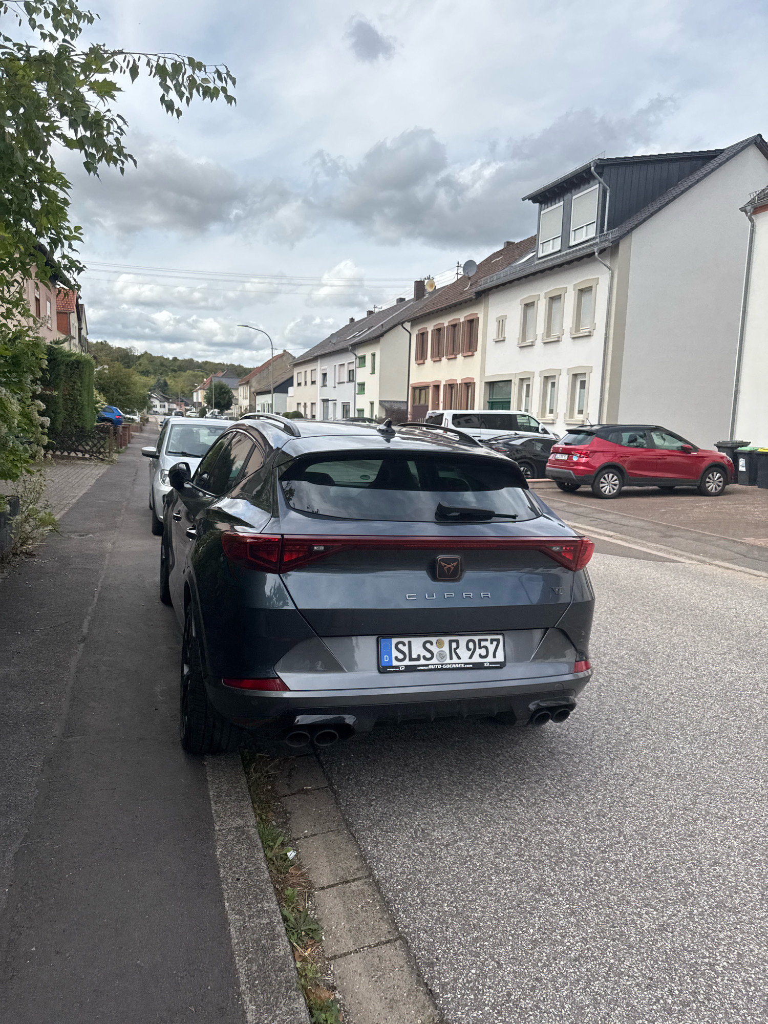 This is a photograph of a street in Überherrn, Germany, on a cloudy day. The image is taken from a low angle, focusing on the rear of a dark grey CUPRA SUV parked on the side of the road. The car has a license plate that reads "SLS R 957". Two other cars are visible behind it, one silver and one red. The street is lined with residential buildings, some with multiple stories and dormer windows. Trees and foliage are present on the left side of the street, and a sidewalk is visible in the foreground. The sky is overcast with dramatic cloud formations.