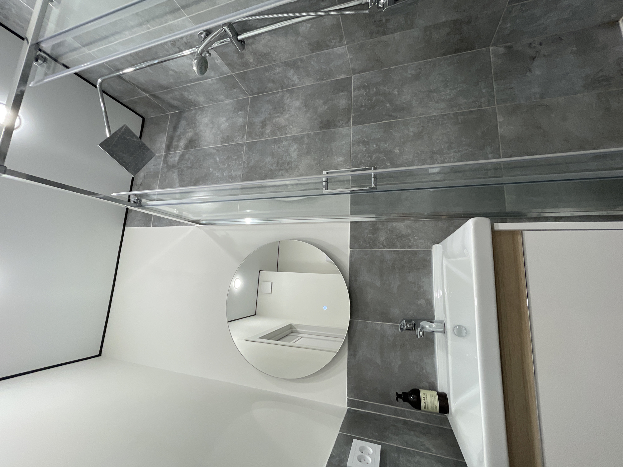The image shows a bathroom interior, likely in Ufa, Russia. The walls are a mix of white paint and gray tiles. A round mirror reflects a bright, possibly adjacent room with a raised platform. A sink with a silver faucet and a dark bottle of liquid soap sits to the right of the mirror. Above, a shower enclosure is visible, with a square shower head and a curved shower curtain rod. The floor appears to be tiled with gray. The image suggests a modern, clean bathroom design.