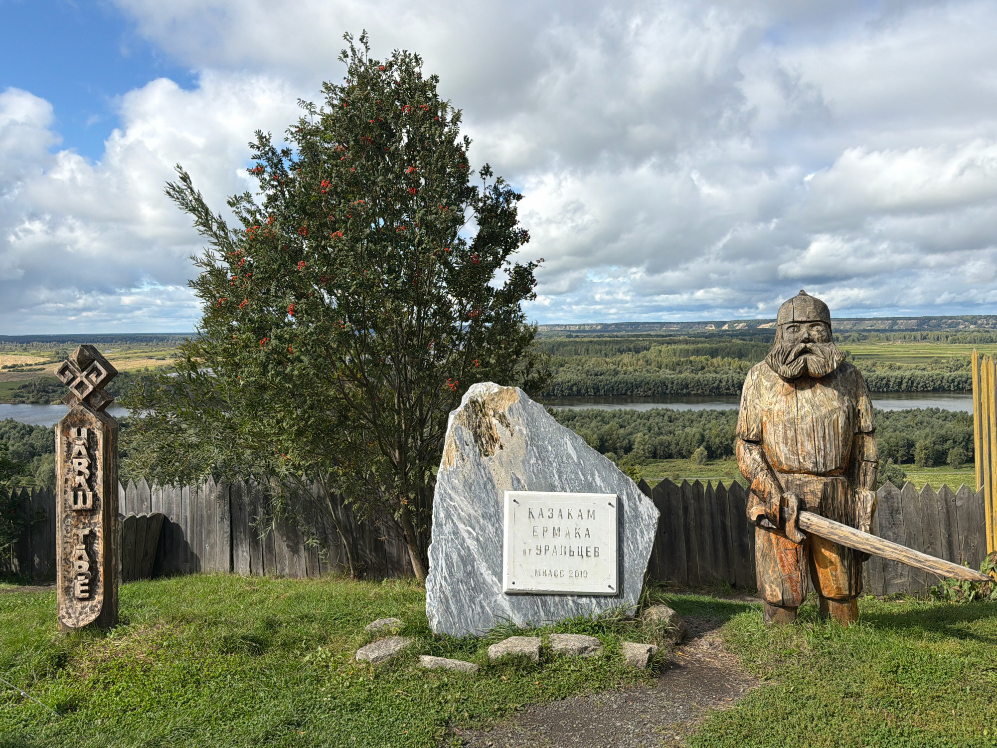 In the city of Абалак, Russia, this image captures an outdoor scene with a historical theme. In the foreground, a wooden statue of a bearded man wearing a helmet and holding a sword stands on a grassy mound. To the left of the statue is a large, rough stone monument with a white plaque inscribed in Russian. Adjacent to the stone monument is a wooden post carved with symbols and text. Behind these elements, a wooden fence encloses the area. A tree with red berries is visible behind the fence. In the background, a river flows through a landscape of lush green forests and distant hills under a cloudy sky. The scene appears to be a memorial or tourist attraction, possibly related to Cossack history.