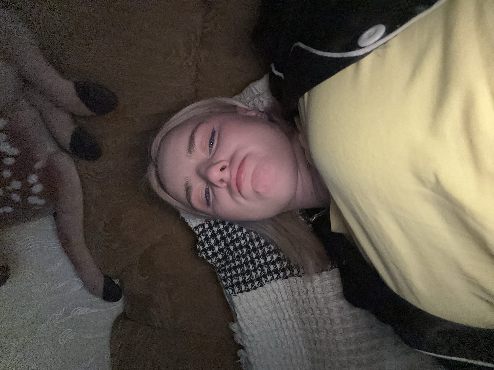 A young woman with blonde hair and blue eyeliner is lying on a couch, taking a selfie. She is wearing a yellow shirt and a black jacket with white stripes. Behind her, there is a plush deer toy. The couch is covered in a brown, textured fabric. A patterned throw blanket is draped over the couch next to her. The lighting is dim, suggesting the photo was taken indoors at night.