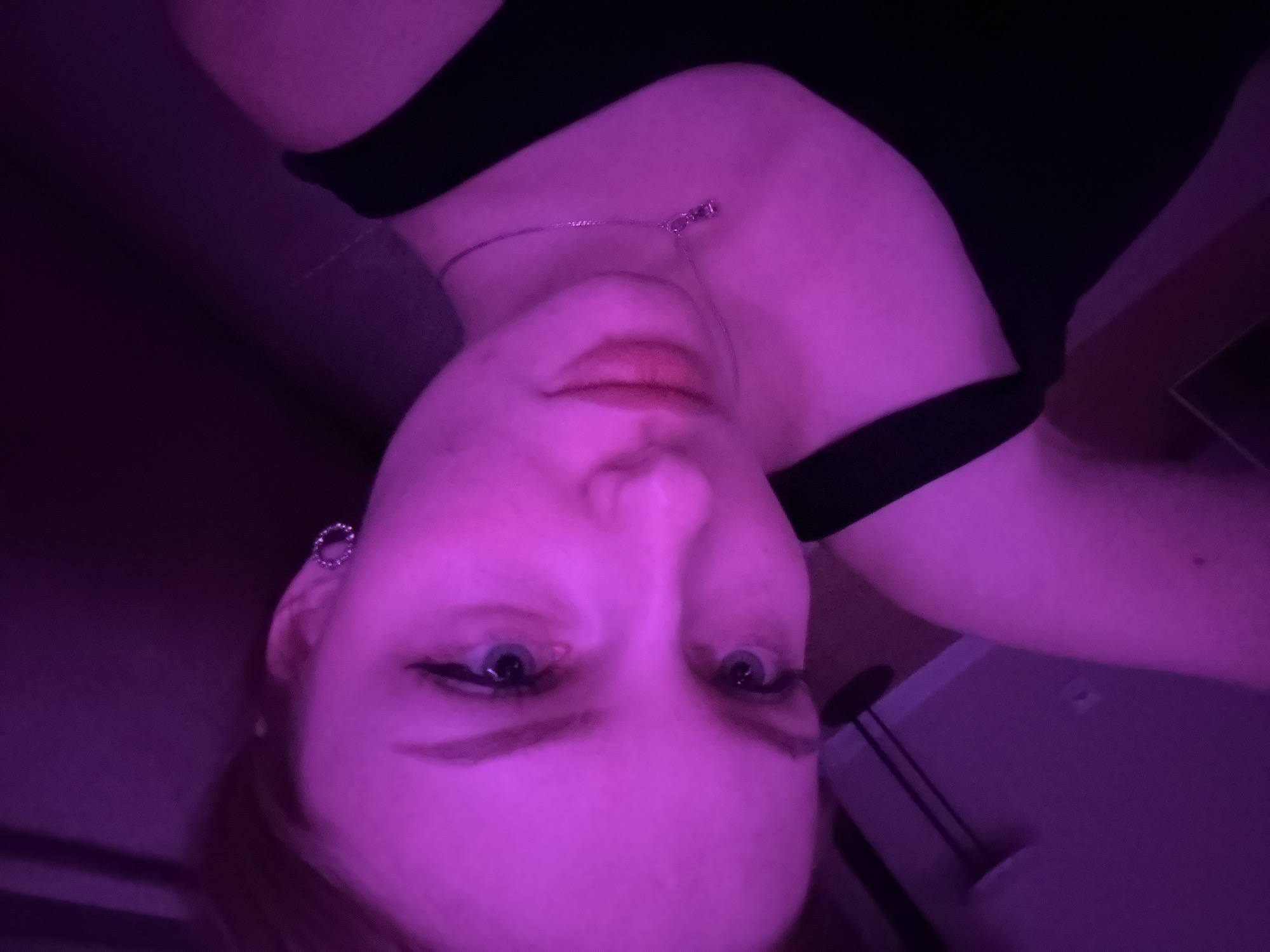 A photograph depicts a woman with fair skin illuminated by a purple light. She is wearing a black garment that exposes her neck and shoulders. A thin silver necklace is visible around her neck. She has blue eyes and dark eyeliner. An earring is present on her left ear. The background is dark and partially obscured, with hints of furniture and what appears to be a computer monitor. The photograph is oriented upside down. There is no information that indicates the location is Perm, Russia.