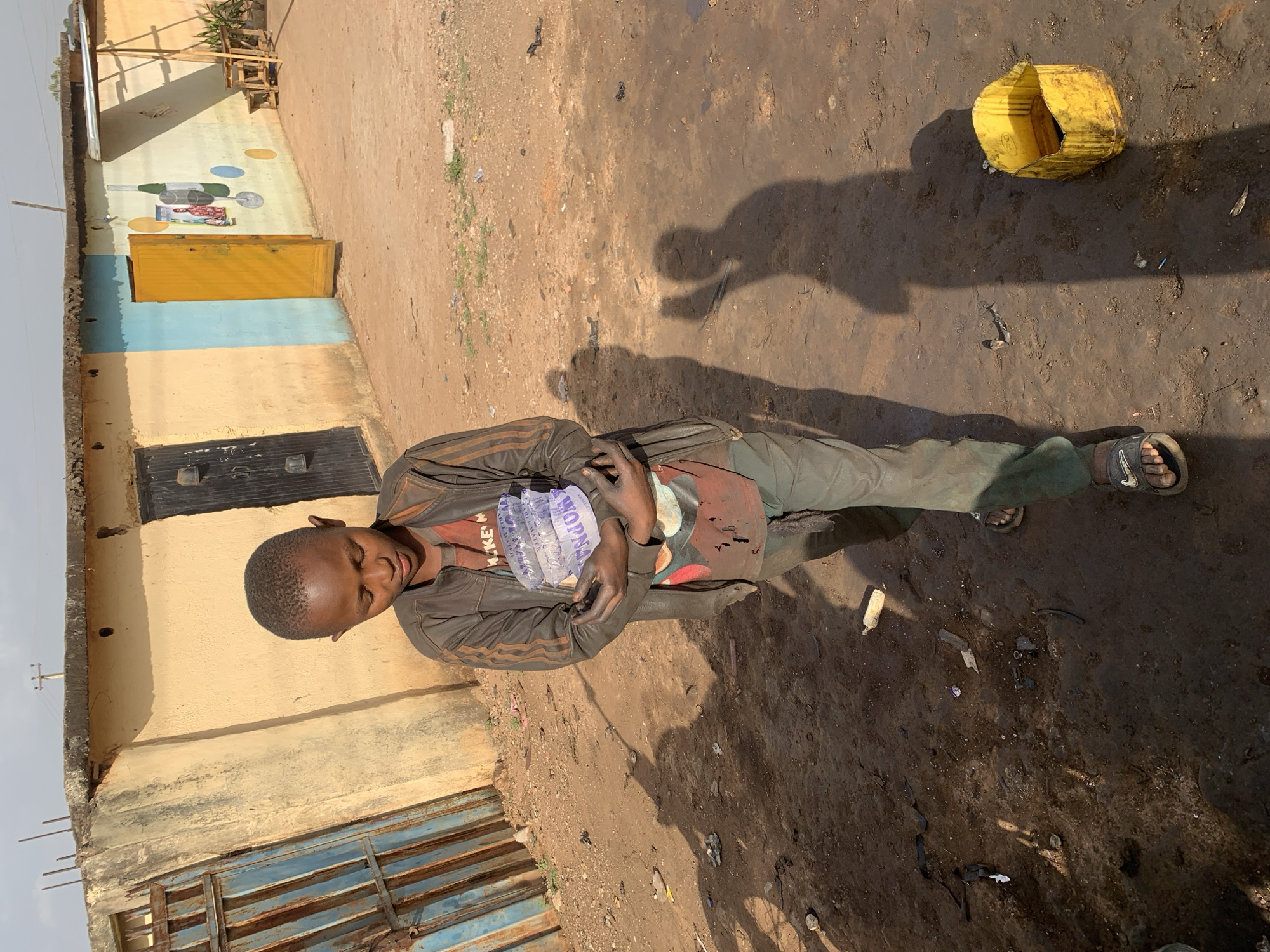 A person is lying on the ground in an outdoor environment. The person is wearing a brown jacket, green pants, and sandals. They are holding a plastic bag filled with water. A yellow plastic container is nearby. The ground is dirt and debris. A building with a blue door and yellow wall is visible in the background. The location is Gura Top, Nigeria.