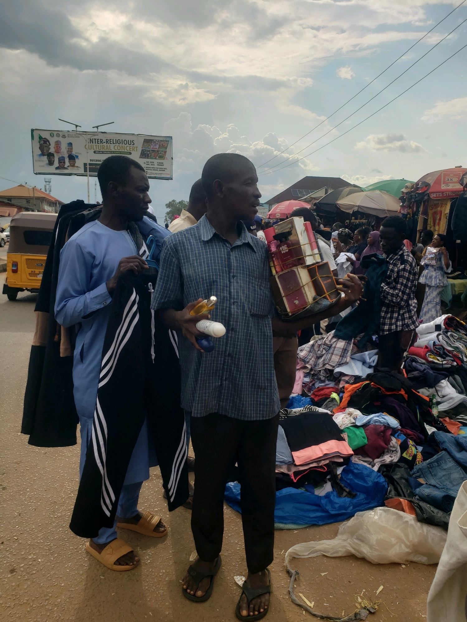 The image depicts a street scene in Sabon Tasha, Nigeria, likely a market area. Two men are the primary subjects in the foreground. The man on the left, wearing a light blue traditional outfit and beige sandals, is holding a dark garment with white stripes on the side. The man on the right, dressed in a blue checkered shirt and dark trousers with black sandals, is holding a stack of boxed items, possibly books or religious texts, and two bottles, one appearing to be shampoo or lotion.
Behind them, a bustling market is visible with stalls covered by umbrellas, displaying various clothing items piled on the ground. A yellow auto-rickshaw (keke napep) is parked on the left side of the road. The sky is overcast with clouds. The overall environment suggests an outdoor marketplace with commercial activity.