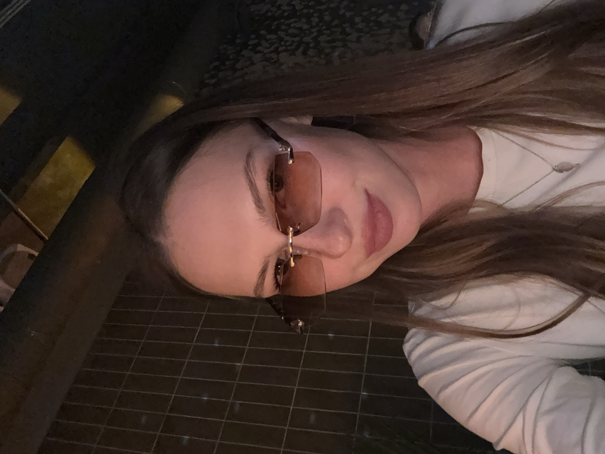 A woman wearing rimless sunglasses with amber lenses is pictured from the chest up. She has long, brown hair and is wearing a white shirt. The background is dark and appears to be an indoor setting with a tiled wall on the left and a textured surface on the right. There is no discernible indication of the specific location being in the city of Perm, Russia.