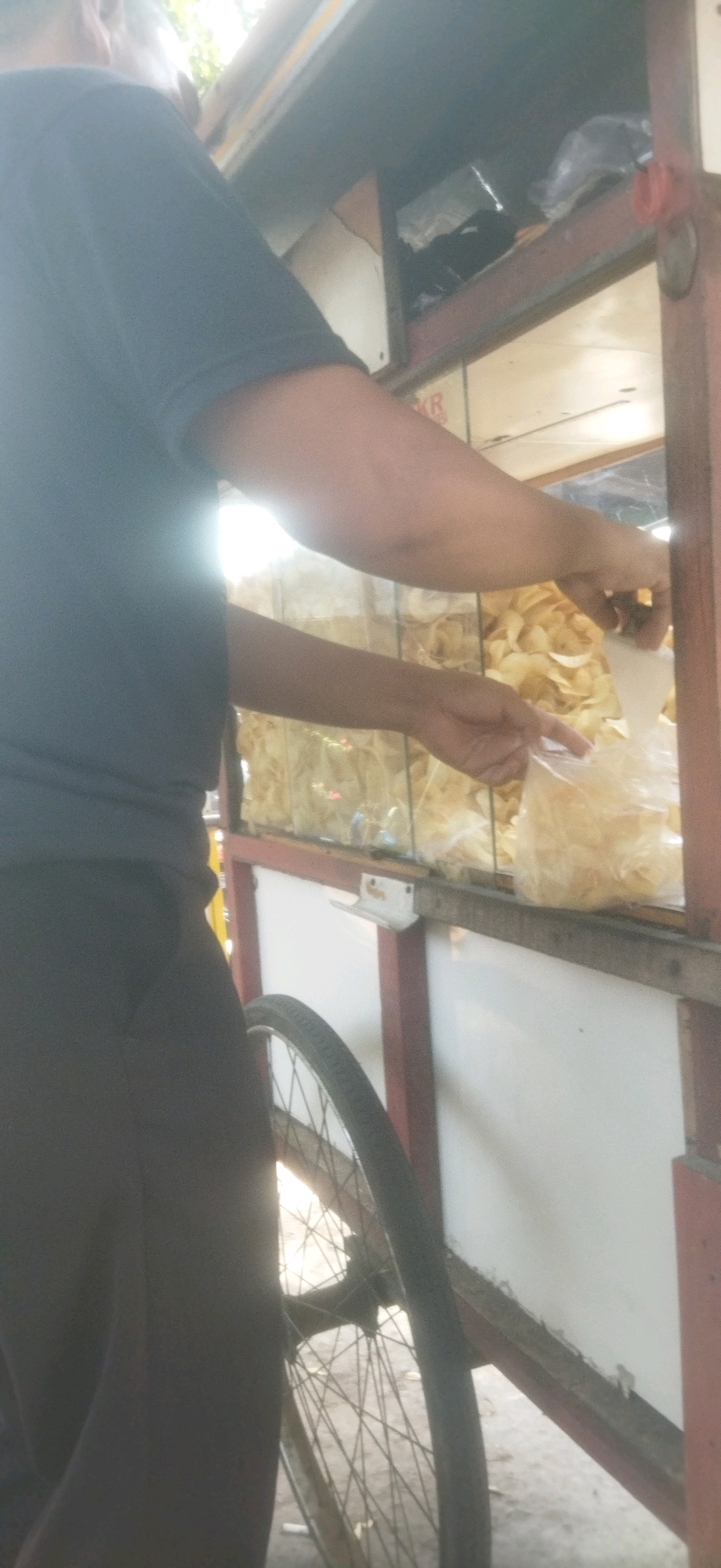 The image shows a close-up view of a person in a dark blue shirt and dark pants, standing next to a food cart. The cart has a glass display case filled with what appear to be packaged snacks or food items. One of the person's hands is reaching into a clear plastic bag containing light-colored, flat food items, possibly chips or crackers. The cart has a large spoked wheel visible on the right side. The background suggests an outdoor setting, with diffused light indicating it is daytime. The person's head and face are not clearly visible. The overall scene depicts a commercial transaction at a street food vendor.