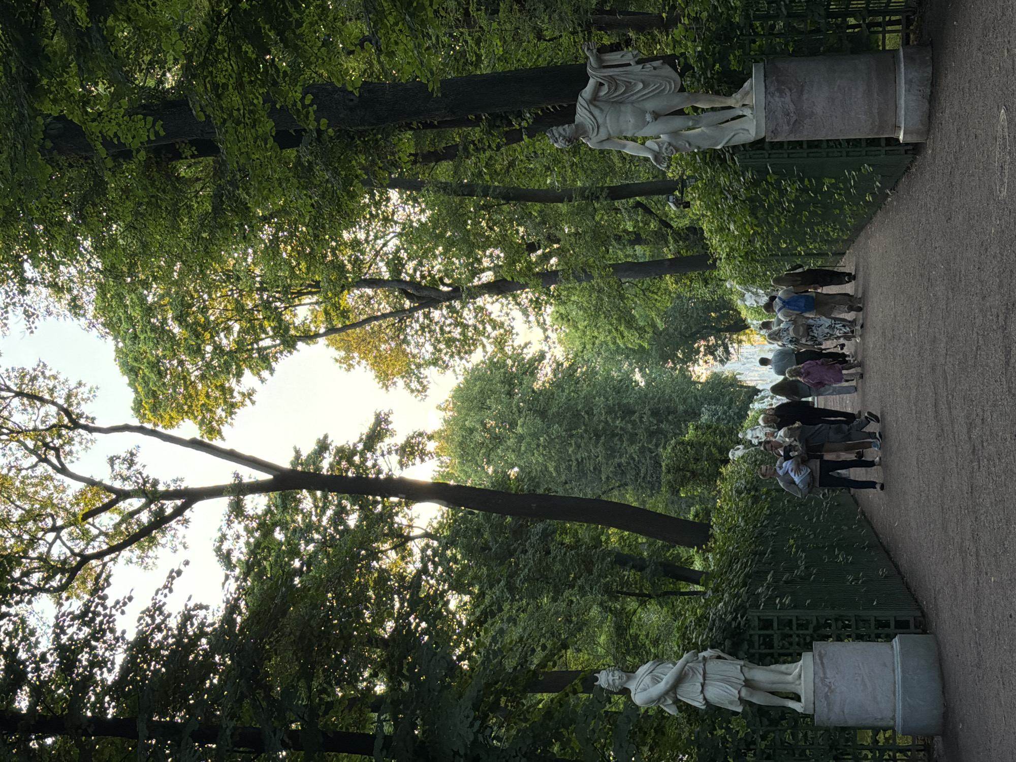 The image depicts a pathway in a park in Saint Petersburg, Russia, lined with lush green trees and ornate sculptures. On the left side, a female statue stands on a pedestal, and on the right, a male statue is similarly displayed. A group of people, including adults and children, are walking along the pathway, appearing to be tourists. The scene is bathed in soft, natural light, suggesting it is either early morning or late afternoon. In the distance, a fountain is visible, adding to the picturesque ambiance of the park. The pathway itself is paved with gravel and bordered by a low green fence. The overall atmosphere is serene and inviting, characteristic of a well-maintained public garden.