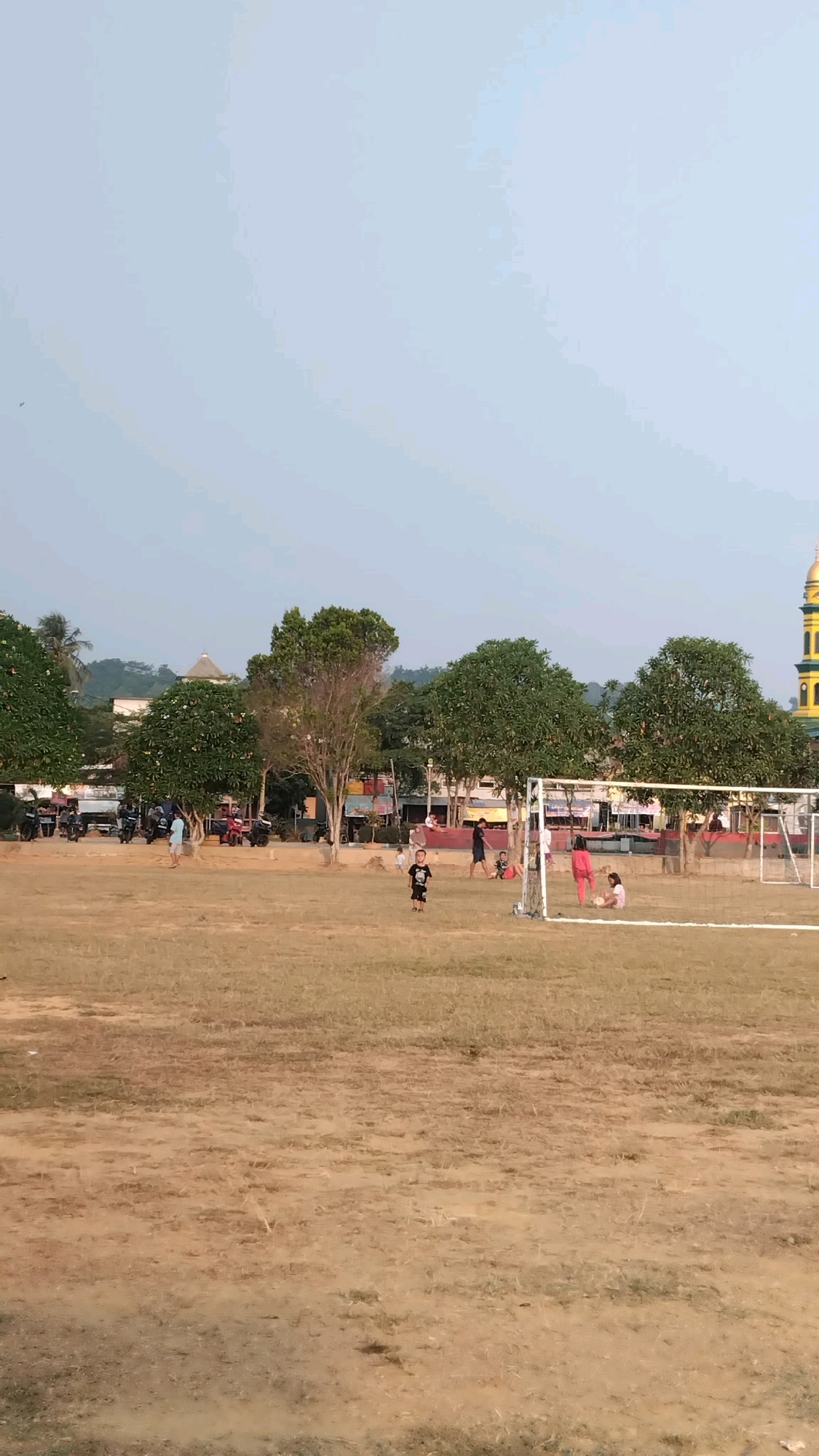 The image depicts a sports field in Manunggal Jaya, Indonesia, characterized by dry, light-brown grass and patches of dirt. In the foreground, a young child in a black t-shirt and shorts stands near the center of the frame. To the right, a soccer goal with a white net is visible, and several individuals, including two children, are near it. One child is wearing a pink shirt and shorts, and another is in a light-colored shirt and shorts.
In the background, a row of trees with lush green foliage forms a visual boundary. Beyond the trees, buildings are visible, including a structure with a prominent gold dome and a tall minaret, likely a mosque. Several motorcycles are parked in front of some buildings. The sky is clear and light blue, suggesting a daytime scene. The overall impression is that of a community space being used for recreation.