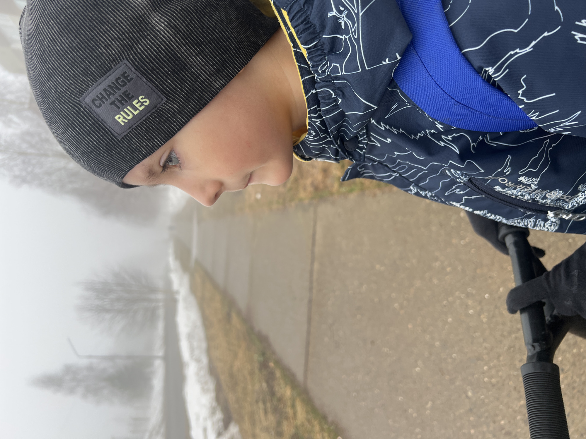 A child wearing a dark gray beanie with a "CHANGE THE RULES" patch and a dark blue jacket with white drawings and a blue backpack strap is riding a scooter along a sidewalk in what appears to be a foggy or misty environment. The sidewalk is next to a grassy area with snow on the ground. In the background, there are trees partially obscured by the fog. The location is Марковский, Russia, inferred based on the picture's metadata.