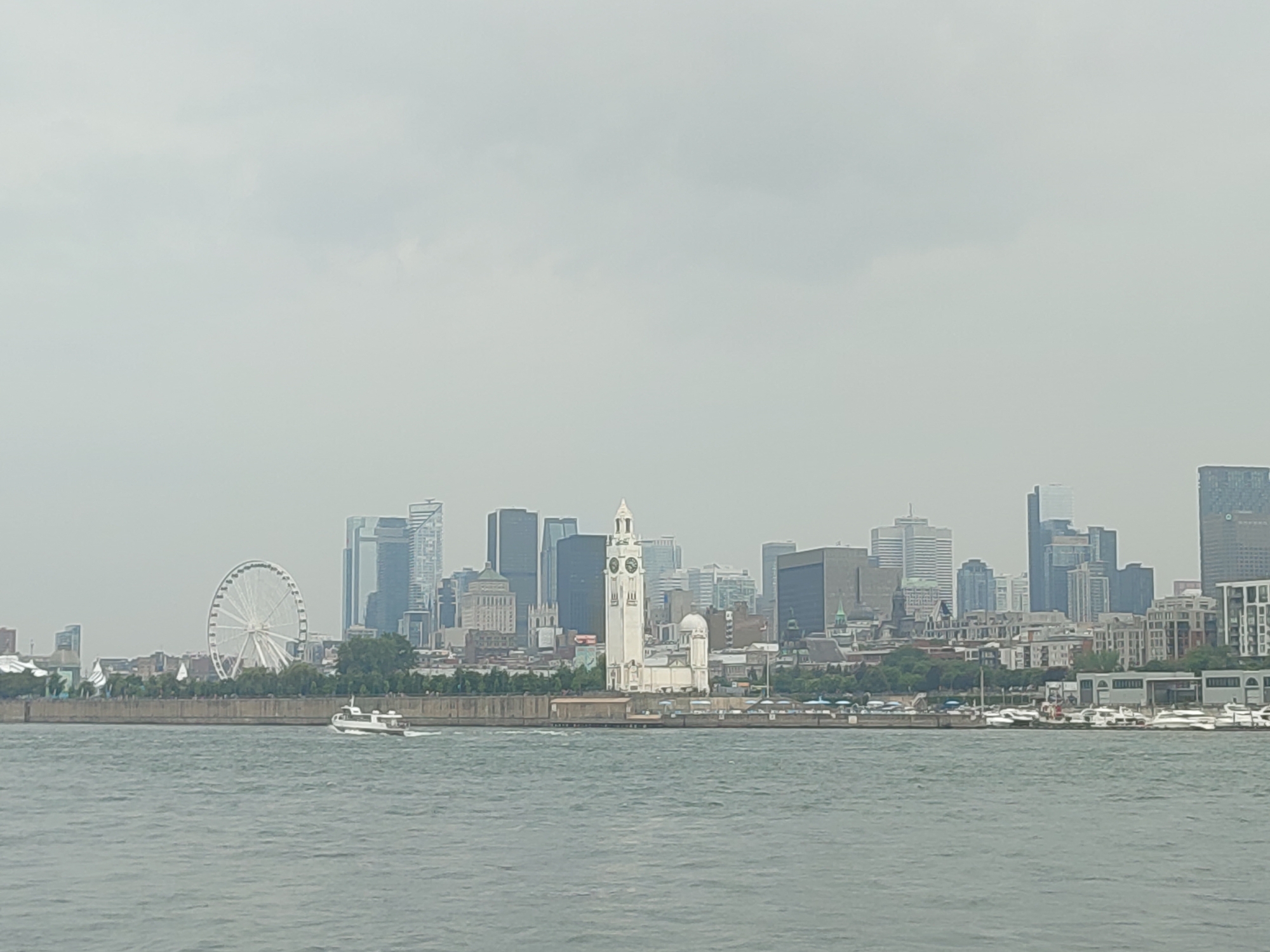 The media file depicts a panoramic view of the Montreal skyline from across a body of water. The skyline is dominated by a collection of modern skyscrapers, with a prominent white clock tower featuring a dome and multiple clock faces standing out in the foreground. To the left of the clock tower, a large Ferris wheel is visible, suggesting a recreational area. The water in the foreground is choppy and grey, with a white boat moving from left to right. Along the waterfront, there's a line of buildings and trees, and further down the shore, a cluster of white yachts are docked. The sky is overcast and a hazy grey, indicating a cloudy or possibly foggy day. The overall impression is of a bustling urban environment observed from a distance, with a muted color palette due to the weather. No specific time of day is explicitly discernible beyond it being daytime, and no people are clearly visible in the image. The location cues of the Ferris wheel and the distinctive clock tower strongly point to Montreal, Canada.