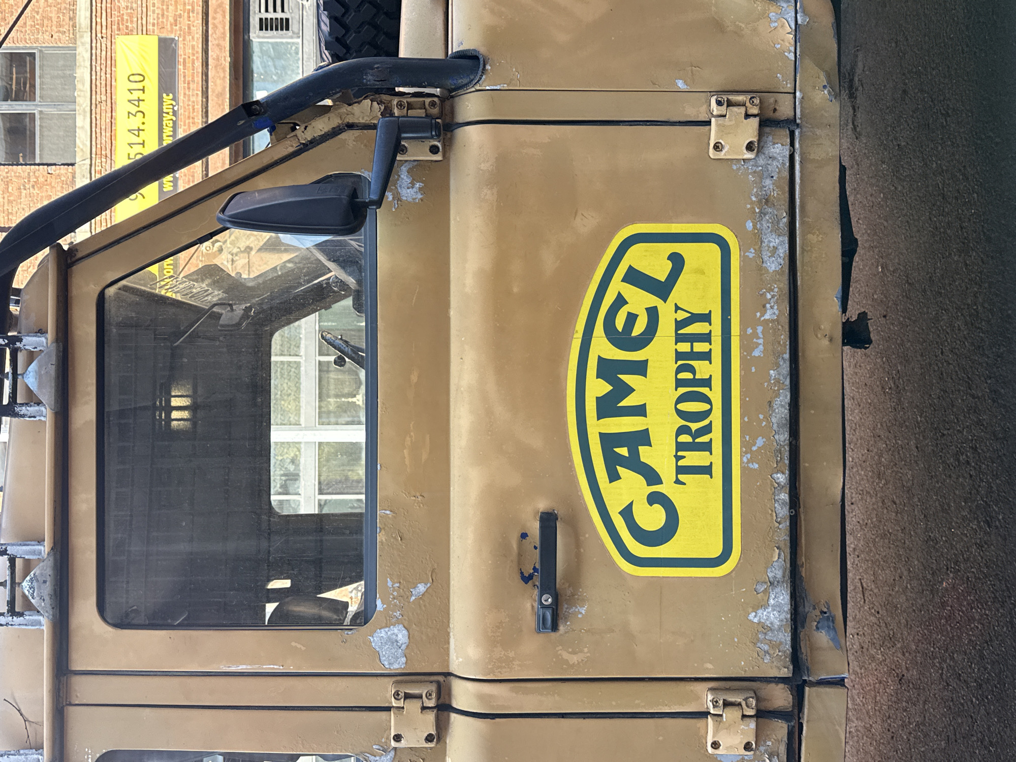 The image depicts the side of a Camel Trophy-branded Land Rover, likely a vintage vehicle known for its participation in the off-road competition. The vehicle is painted in a faded yellow color with signs of wear and tear. The Camel Trophy logo is prominently displayed on the side. The vehicle's window, mirror, and door hardware are visible.
In the background, a building with a banner that reads "914.3410 www.rway.nyc" is visible. This banner suggests that the location may be in or near New York City, as the area code 914 is commonly associated with Westchester County, New York, which is near New York City. The image provides the most detail about the vehicle itself, indicating that it may be the focal point of the scene.