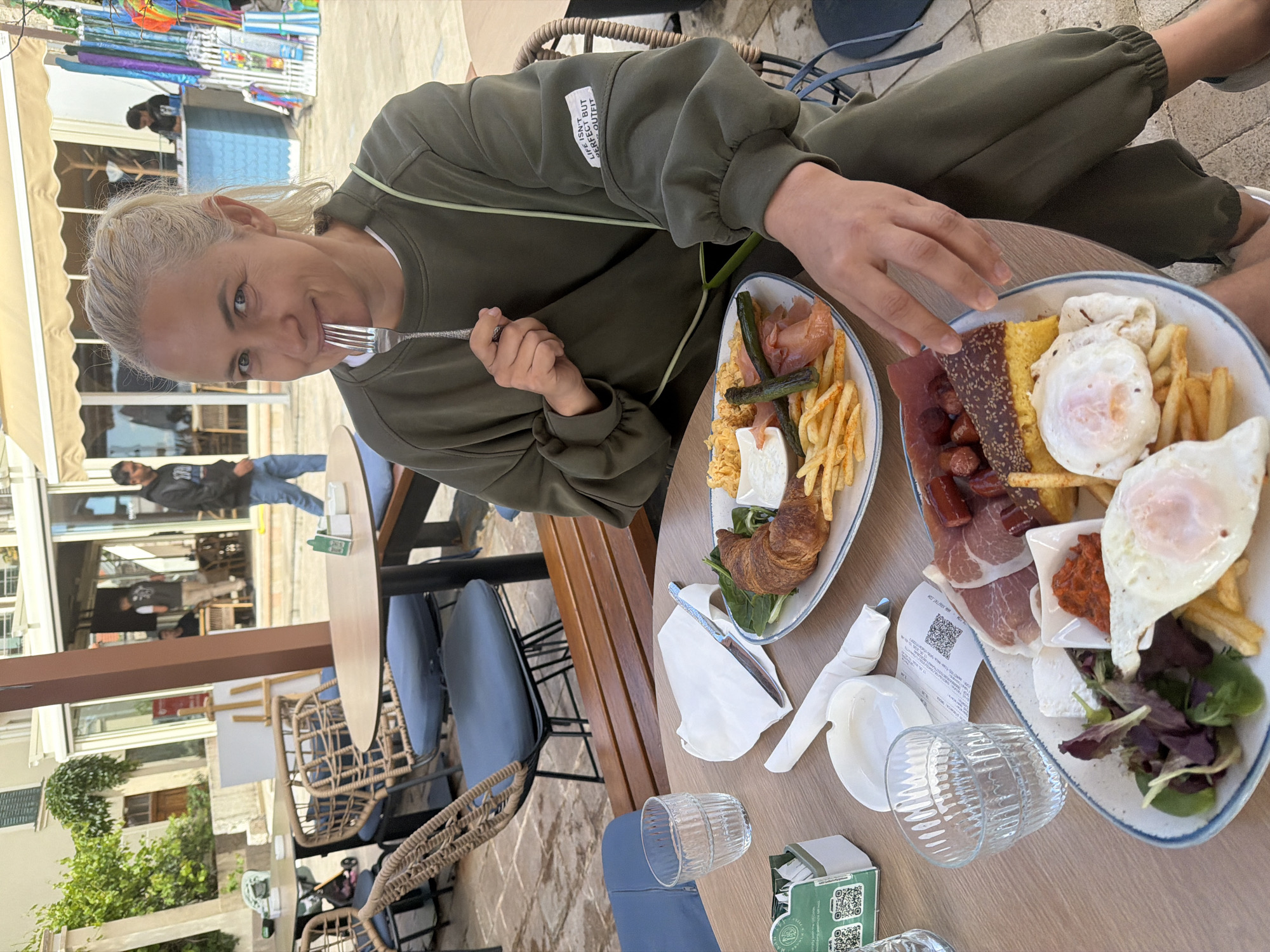 A seated person is shown at an outdoor dining table. She is fair-skinned with light hair. She wears a green sweat suit. She is eating food off a plate with a fork. On the table are two plates of food that contain items like eggs, French fries, meat and vegetables. Also on the table are glasses, napkins, a receipt, and a butter dish. The table is surrounded by blue chairs and a woven wicker chair. The location is the city of Petrovac centar, Montenegro.