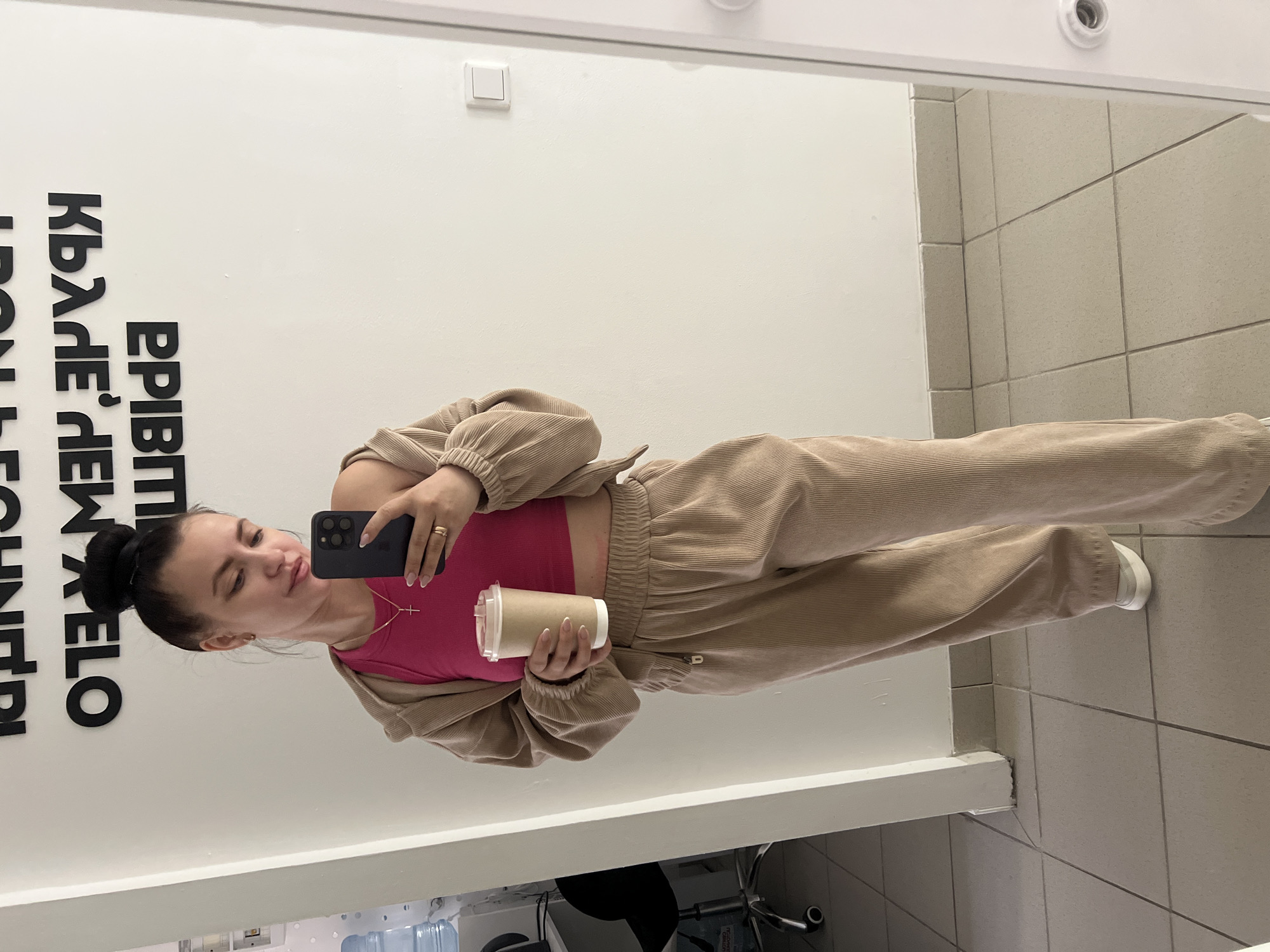 A young woman with her hair pulled up in a bun is taking a mirror selfie. She is wearing a pink crop top, a beige corduroy jacket, and matching beige corduroy pants. She is holding a disposable coffee cup in her left hand and her right hand is resting on her hip. She is wearing a gold cross necklace and a gold ring on her right hand. The background is a white wall with black text on it, a white door frame, and a tiled floor. The overall impression is one of casual style and personal photography. The text on the wall, which is mirrored, appears to be in Russian.
