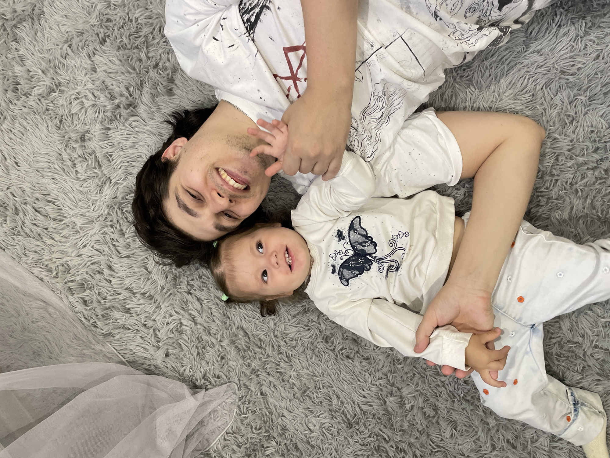 A man and a young child lie together on a gray shag rug. The man has dark hair and is smiling. He is wearing a white patterned shirt. The child, wearing a white shirt with a butterfly design and white pants with small orange dots, is also smiling. The man has his arms around the child. A piece of white tulle fabric is visible in the lower left corner of the image. The location is likely an indoor space, possibly a home, but the scene does not depict any visual features that can be used to conclusively say it is in Izhevsk, Russia.