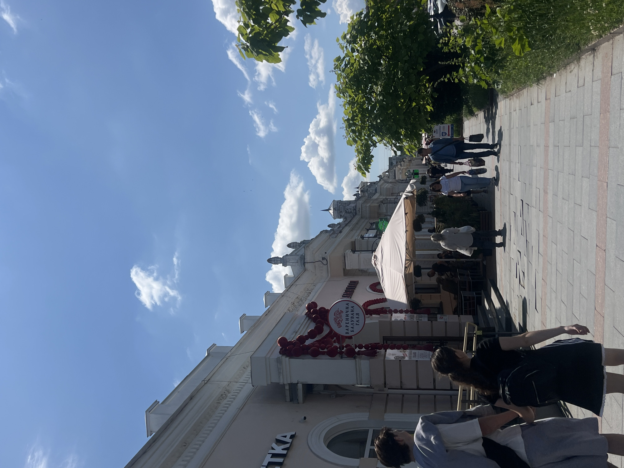 A wide-angle, eye-level outdoor shot captures a sunny day on a European-style pedestrian street in Unknown, Unknown. A light pink building with ornate architectural details lines the left side of the street, featuring arched windows and decorative trim. A sign with red lettering hangs from the building, accompanied by red balloon decorations. Further down the street, awnings of cafes and shops extend onto the sidewalk. A lush green tree with dense foliage is situated on the right side of the scene, with its branches reaching towards the bright blue sky dotted with scattered white clouds. The sidewalk is paved with large, grey interlocking stones and bordered by a strip of reddish-pink paving stones. Several people are walking on the sidewalk. In the foreground, a young couple walks away from the camera, holding hands; the male is wearing a grey hoodie and shorts, and the female is wearing a black dress and a backpack. Other individuals are scattered along the sidewalk, some walking, some appearing to be patrons of the establishments. The overall impression is one of a vibrant and pleasant urban environment on a clear day.