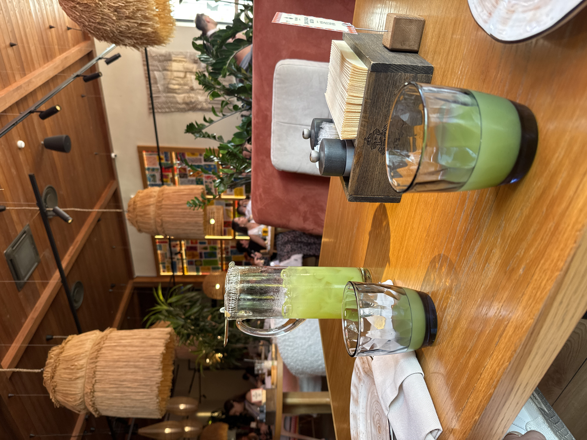 The image shows a close-up view of a table in a restaurant, with two glasses of green juice and a pitcher of the same drink. The table is made of wood and has a light brown color. In the background, there are other tables, chairs, and people dining. The restaurant has a rustic and bohemian decor, with wooden ceiling beams and lamps made of natural materials. The overall lighting is warm and inviting. The scene is set in the city of Estosadok, Russia, as indicated by the provided context. The image is captured from a low angle, focusing on the drinks and the table setting. The atmosphere appears relaxed and convivial.