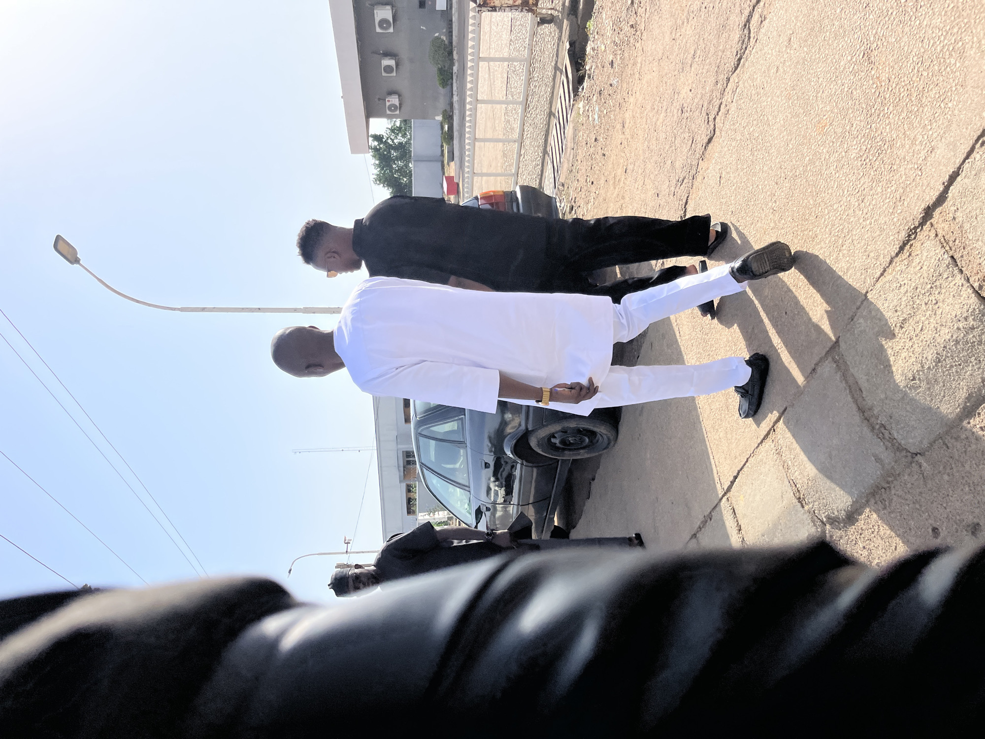 In Bukuru, Nigeria, a scene unfolds on a sunny day, with two men walking on a paved sidewalk. The man on the left, with a shaved head, is dressed in a white traditional outfit and black formal shoes, his posture suggesting he is walking forward. To his right, another man, also with a shaved head and wearing sunglasses, is clad in a black traditional outfit and sandals, walking in the same direction. Behind them, a dark-colored sedan is parked. In the foreground, the blurred image of a person in black clothing is partially visible, adding a sense of depth and perspective. The environment includes a street lamp, power lines overhead, and buildings in the background, suggesting an urban setting. The ground is a mix of dirt and paving stones, indicative of a developing area.