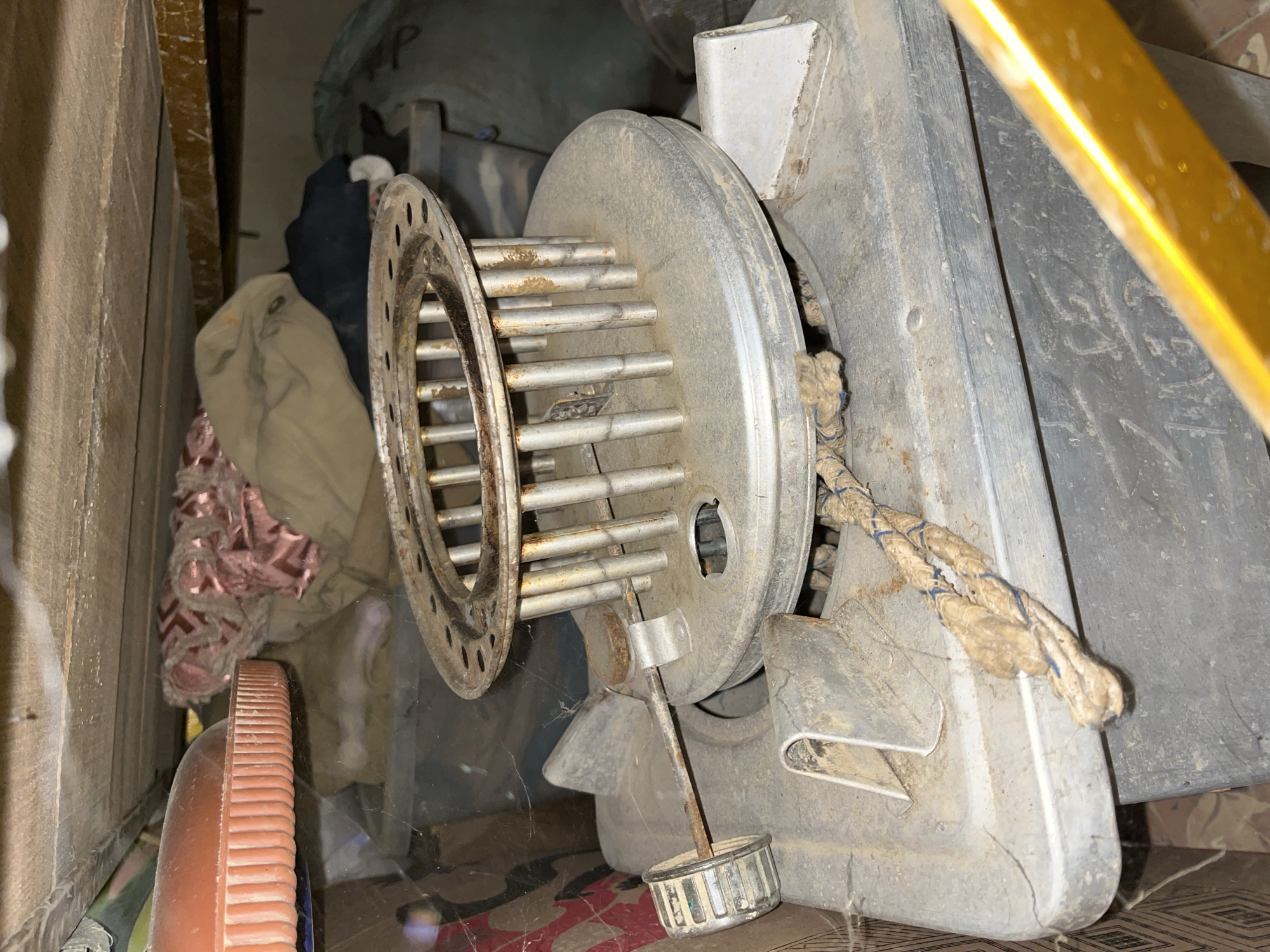 The image shows a close-up view of a dusty, old kerosene stove, possibly a heater, located in a dimly lit storage area. The stove is made of grey metal and has a circular grate with vertical rods on top, which is stained with rust and grime. A thick, frayed rope is attached to the side of the stove. To the left, a control knob with a ribbed surface is visible, connected to a metal rod. The stove sits on top of a dark grey plastic container. In the background, a collection of items is piled up, including what appears to be folded fabric, a brown cloth bag, and other indistinct objects. The storage area has wooden planks forming the ceiling. The overall impression is one of disuse and neglect. The location is described as Kota Kuala Simpang, Indonesia, though the image itself provides no specific visual cues to confirm this location.