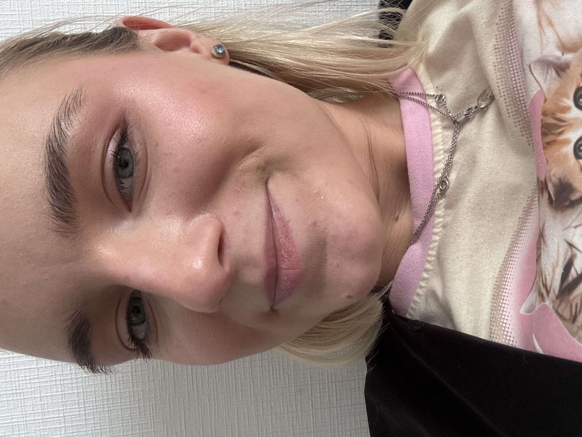 A close-up, tilted selfie depicts a fair-skinned individual with blonde hair, blue eyes, and defined eyebrows. The person is wearing a light-colored shirt with a pink collar and a kitten graphic visible on the fabric. A silver chain necklace is around their neck. An earring is visible in their left ear. The background is a plain white wall. The city and country of Unknown, Russia are not discernible from the image.