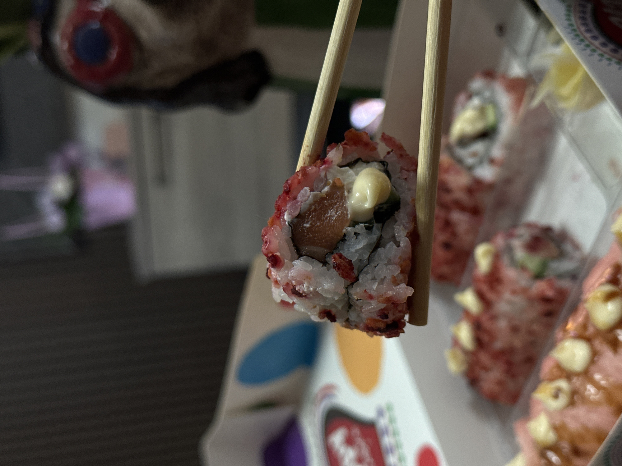 The image presents a close-up, focused shot of a sushi roll held aloft by light-colored wooden chopsticks. The sushi roll is an inside-out (uramaki) type, with white rice on the exterior generously coated in a vivid red, granular topping, likely tobiko (flying fish roe). Visible within the roll's center are a piece of reddish-brown fish, a slice of green vegetable (possibly cucumber or avocado), and a dollop of creamy white or light yellow sauce.
In the blurred background to the right, several other sushi rolls are visible within a white takeout container, which also features colorful, abstract designs on its side. These background rolls appear to have similar red toppings and creamy accents. To the upper left, a highly blurred, dark object with a distinct red and blue circular feature is faintly discernible, possibly a toy or decorative item. The overall setting is indoors, with ambient, artificial lighting suggested by the soft focus and muted background tones. The scene depicts the act of someone about to eat a piece of sushi. No specific text is legible on the container or elsewhere. This event is occurring in Sofia, Bulgaria.