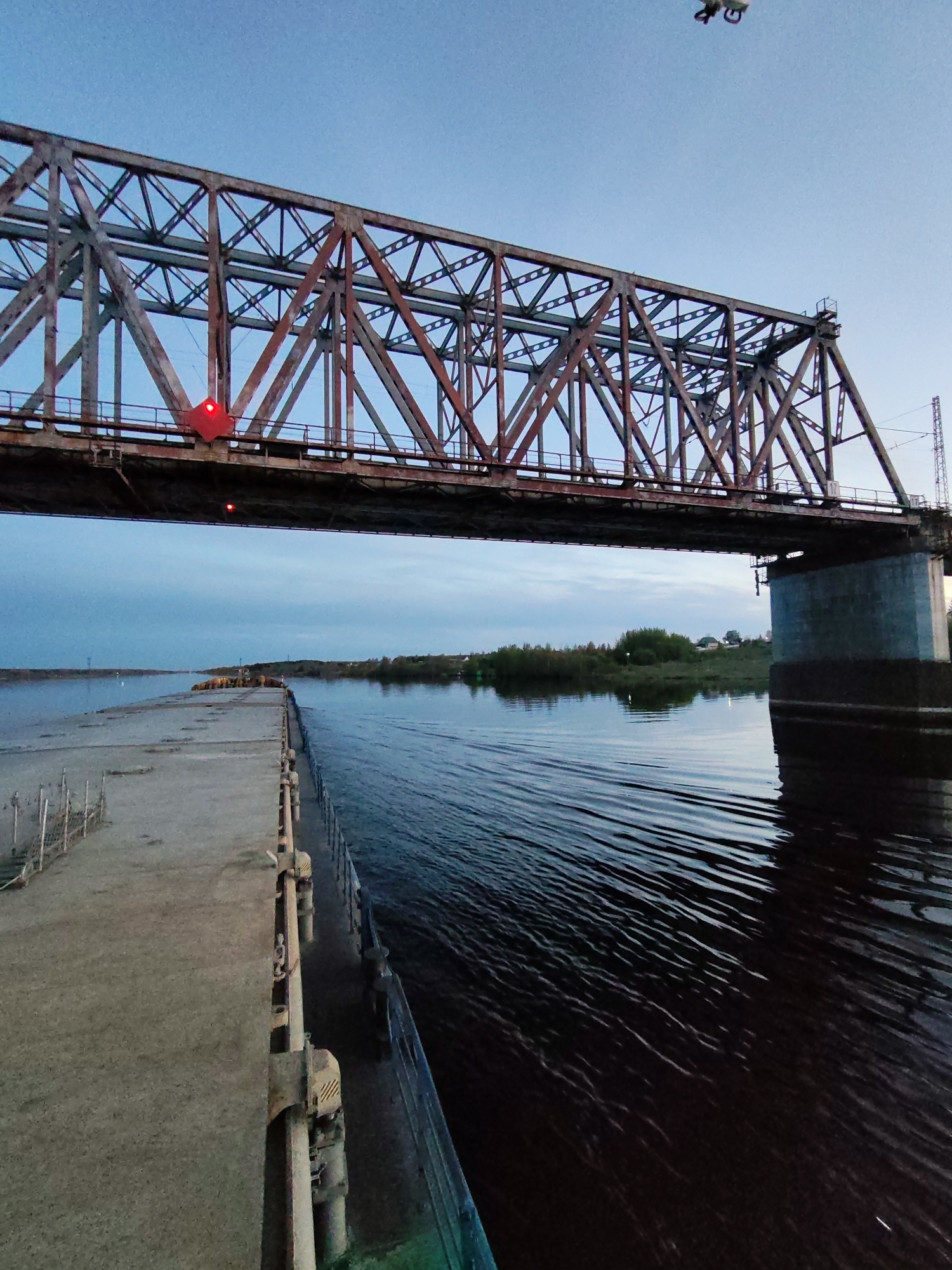The image depicts a steel truss bridge spanning a river, likely in Tarkanovo, Russia. The bridge has a reddish-brown, weathered appearance with a complex lattice structure. A concrete pier supports one end of the bridge on the right side. Underneath the bridge, on the left, there's a concrete dock or platform extending into the river. The water appears dark and slightly choppy, with visible ripples and reflections. The opposite riverbank is visible in the distance, featuring trees and some houses. The sky is overcast, with a gradient of light blue to gray, indicating either dawn or dusk. A drone is visible near the bridge in the sky. Red lights are attached to the bridge structure.