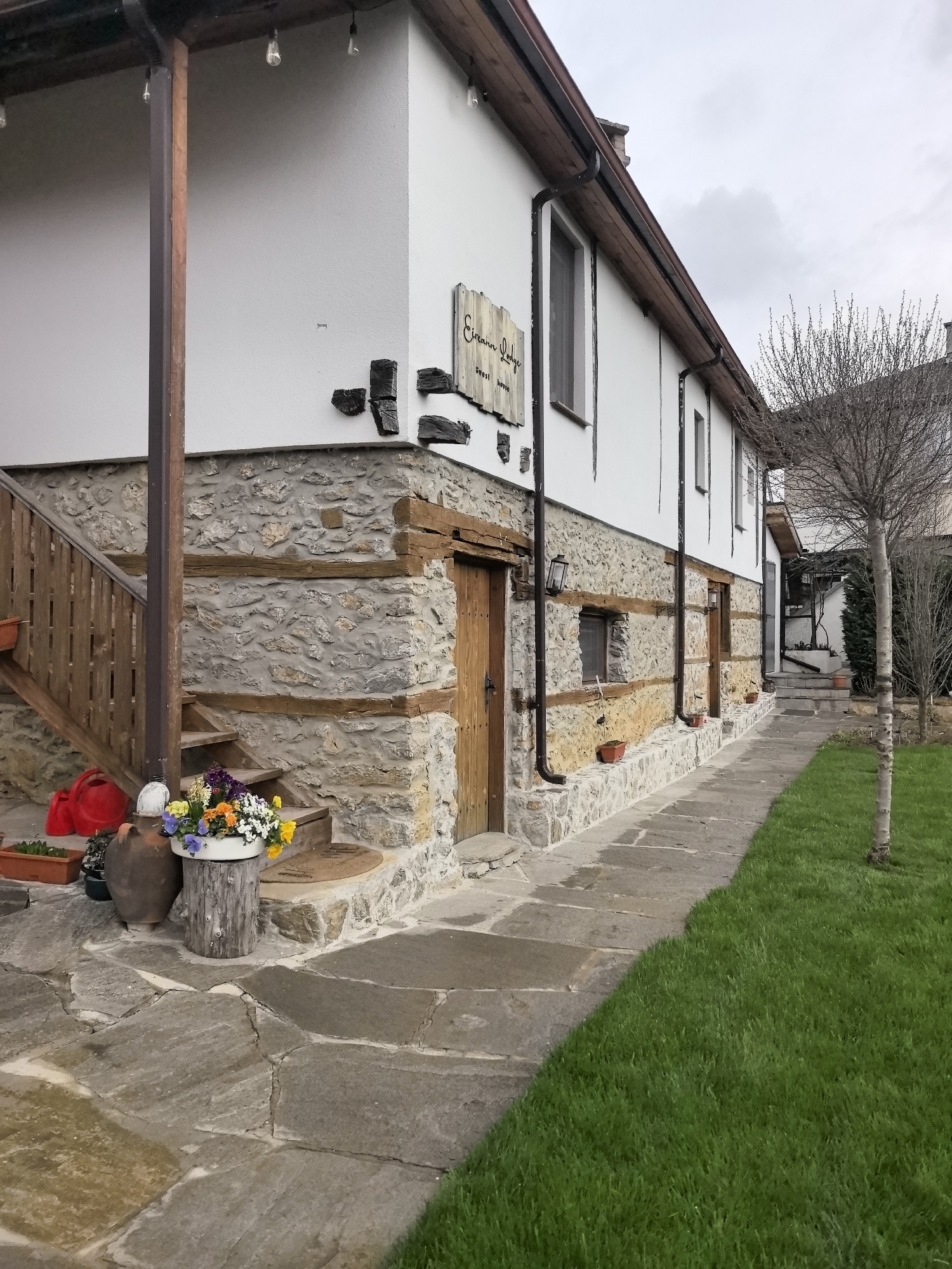 A three-story building with a stone facade is visible. The lower part of the building is constructed of gray stone with wooden beams. The upper floors are white with a dark wooden roof. The building features multiple wooden doors and a few small windows. A sign that reads "Ercann Lodge Guest House" is affixed to the side of the building. A stone pathway leads along the building, bordered by a strip of green grass and a bare tree. A wooden staircase leads up to a veranda on the left side of the building. The location is in an unknown location.