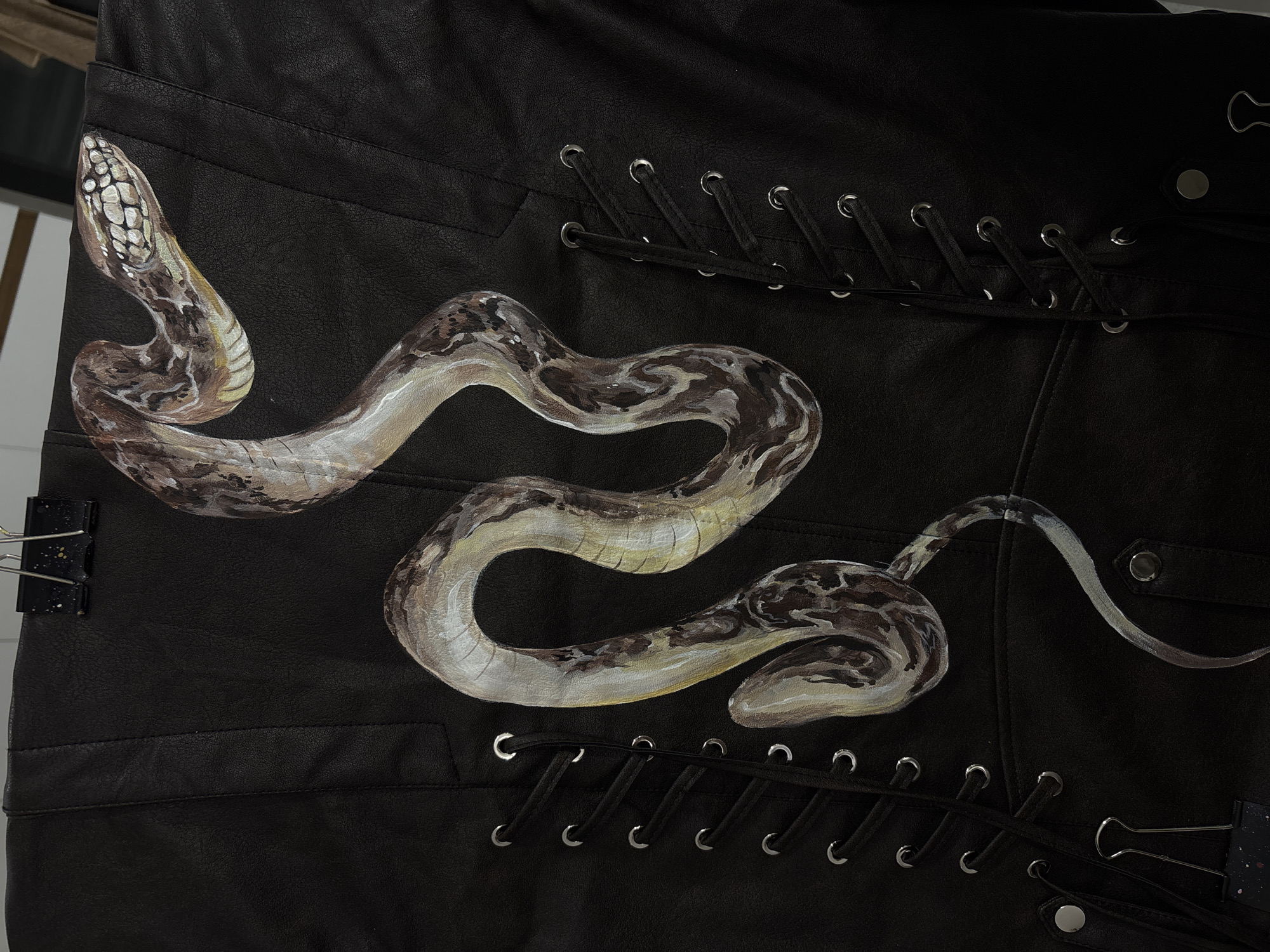 The image shows a close-up of a section of a black leather garment, possibly pants or a jacket. A painted snake, depicted in a realistic style with brown, grey, and white scales, meanders across the leather surface. Metal eyelets are visible along the edges of the garment, laced with black cords. A metal clip is attached to the left side of the fabric. The setting is unknown and does not appear to be specifically Krasnoyarsk, Russia.