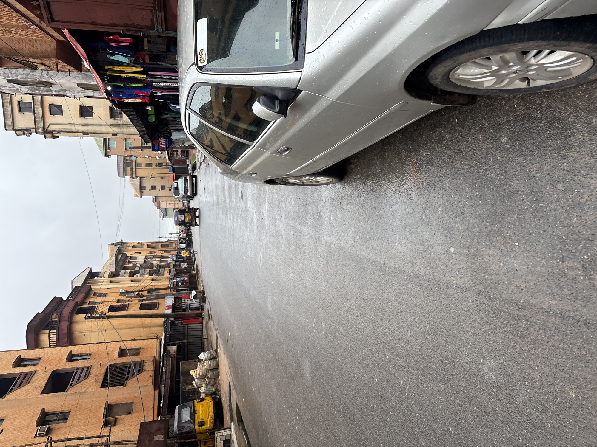 The image depicts a street scene in Onitsha, Nigeria, on an overcast day. The wet asphalt road reflects the gray sky. On the left side of the street are several multi-story buildings with orange and yellow facades. The buildings have balconies and windows, and some appear to be residential or commercial structures with shops at street level. A yellow auto-rickshaw, also known as a "keke" or "marwa," is parked on the left, along with several sacks of goods.
Further down the street, a silver sedan is parked on the right side, close to the camera, its surface speckled with water droplets. Beyond the sedan, the street continues, lined with more buildings and shops. Several auto-rickshaws and a white car are visible in the distance, moving along the road. A few individuals are walking on the sidewalk. The overall impression is of a typical urban environment in Onitsha, likely a market area or a busy thoroughfare.