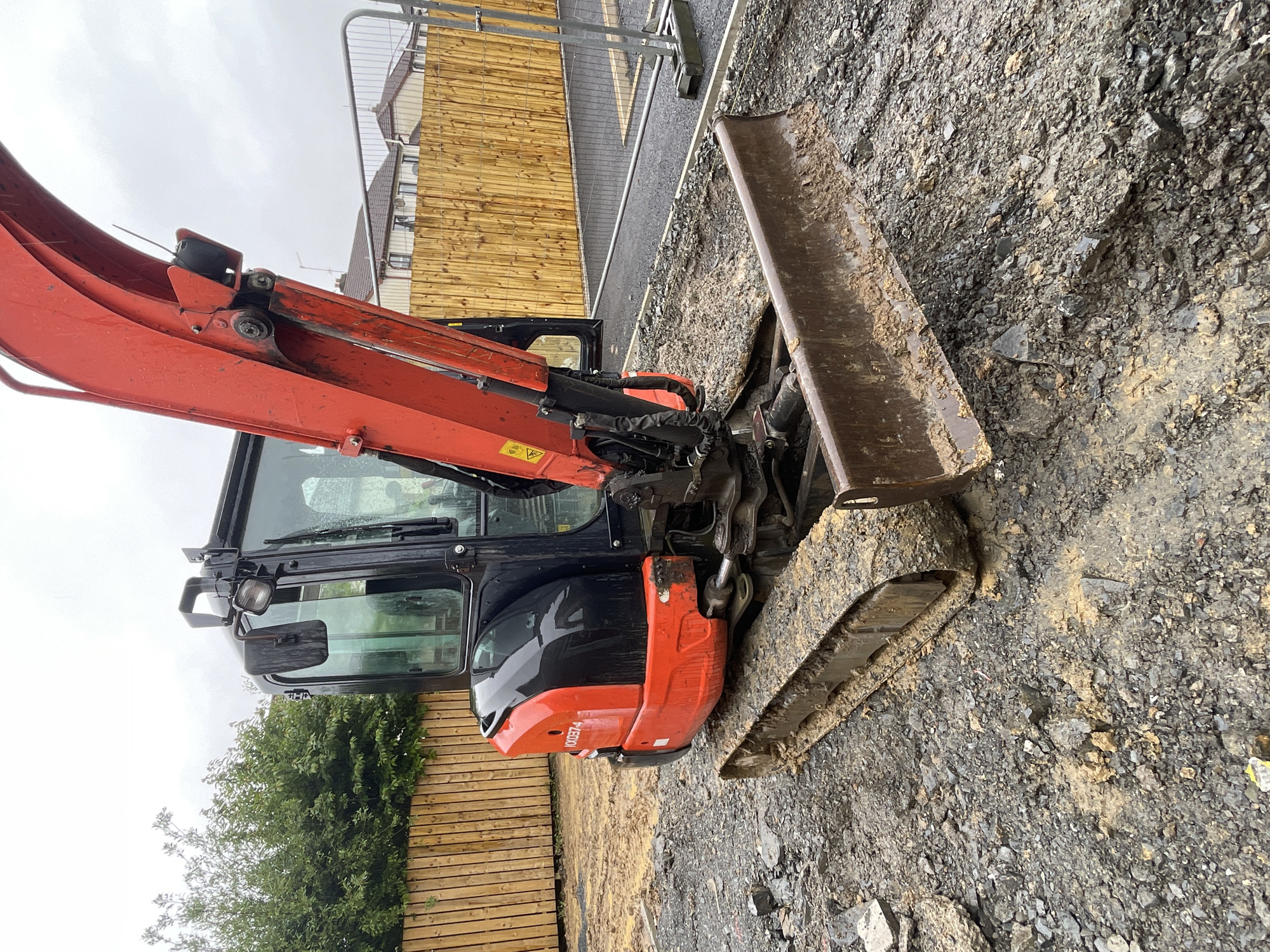 The image shows an orange and black excavator parked on a gravelly construction site. The excavator is the primary subject, with its boom and bucket visible. The background includes a wooden fence, a metal safety barrier, and some buildings that appear to be under construction. The sky is overcast, suggesting cloudy weather.
There are no people visible in the image. The scene is outdoors, likely in a construction or renovation area. The ground is covered in loose stones and dirt, with some muddy patches. The excavator appears to be stationary, with no activity taking place.
Notable details include the model number "U00374" visible on the side of the excavator. The weather appears to be overcast, and the overall impression is one of a working construction site. The location context provided is Banbridge, United Kingdom.