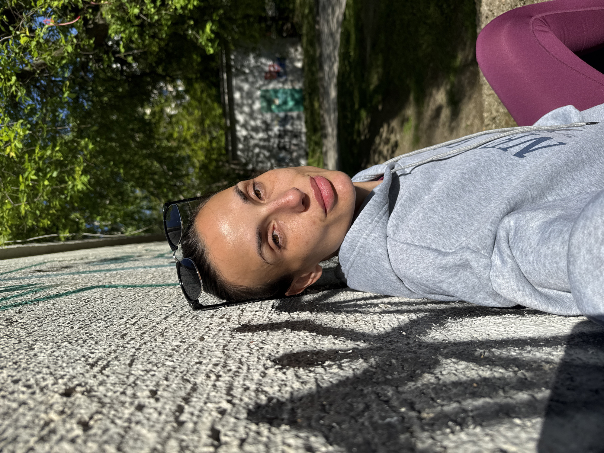 A woman lies on her side on a paved surface outdoors. She is wearing sunglasses, a grey hooded sweatshirt, and maroon leggings. The ground appears to be a textured concrete surface with shadows cast upon it, likely from trees. In the background, trees and a building can be seen. Given the request context of the city of Pleven, Bulgaria, the exact location is unspecified within the image.