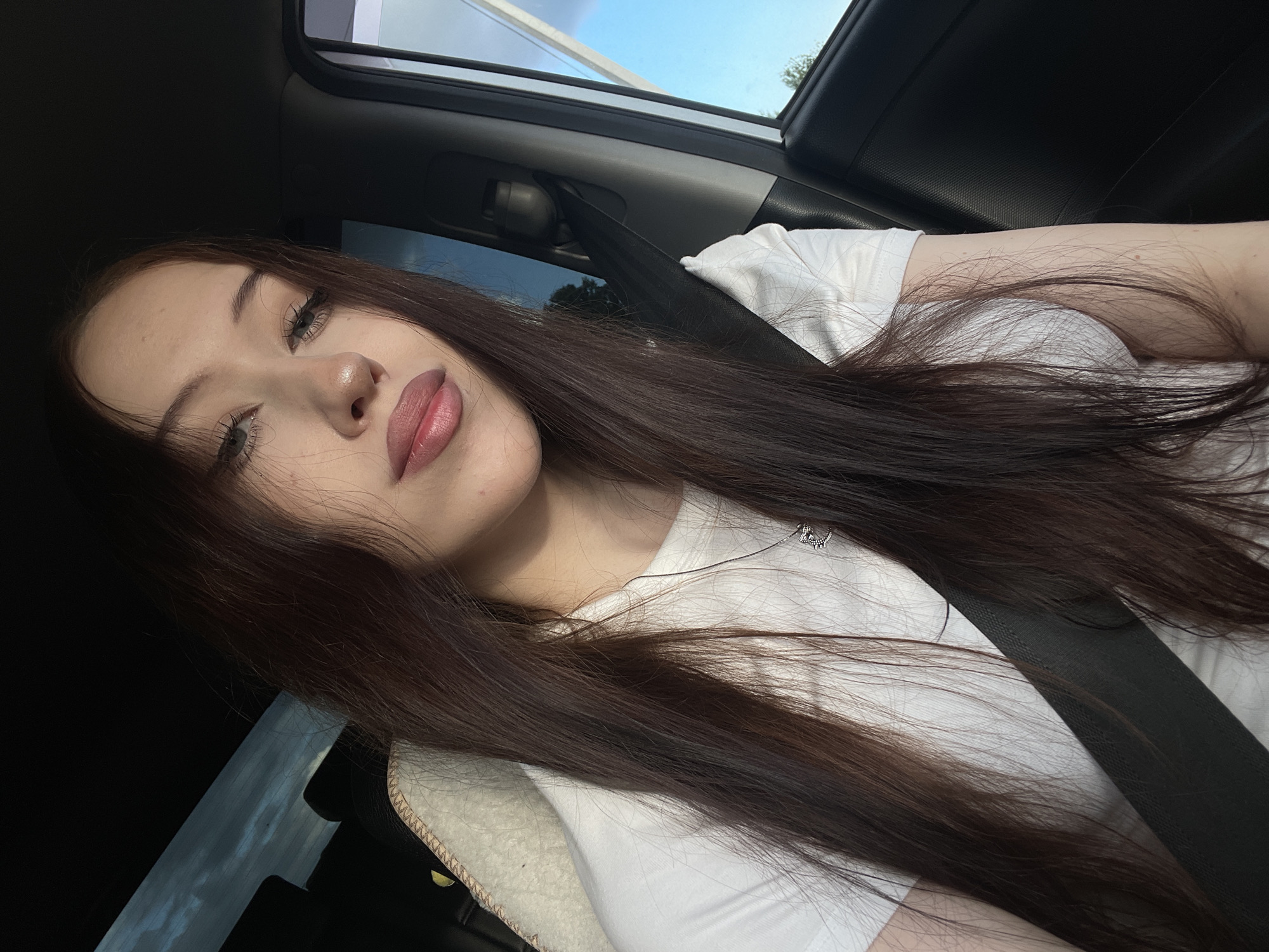 This is a selfie taken inside a car. The subject is a young woman with long, dark brown hair. She is wearing a white t-shirt and a silver necklace with a small charm. Her face is angled towards the camera, with her eyes partially closed and her lips slightly parted. She appears relaxed and is leaning back against the car's headrest, which has a fuzzy, cream-colored trim. A seatbelt is visible across her chest. The background shows the interior of the car, including the window and a glimpse of the sky and trees outside, suggesting it is daytime with clear weather. The lighting suggests it might be late afternoon or early evening due to the warm tones on her skin. There is no visible text in the image.