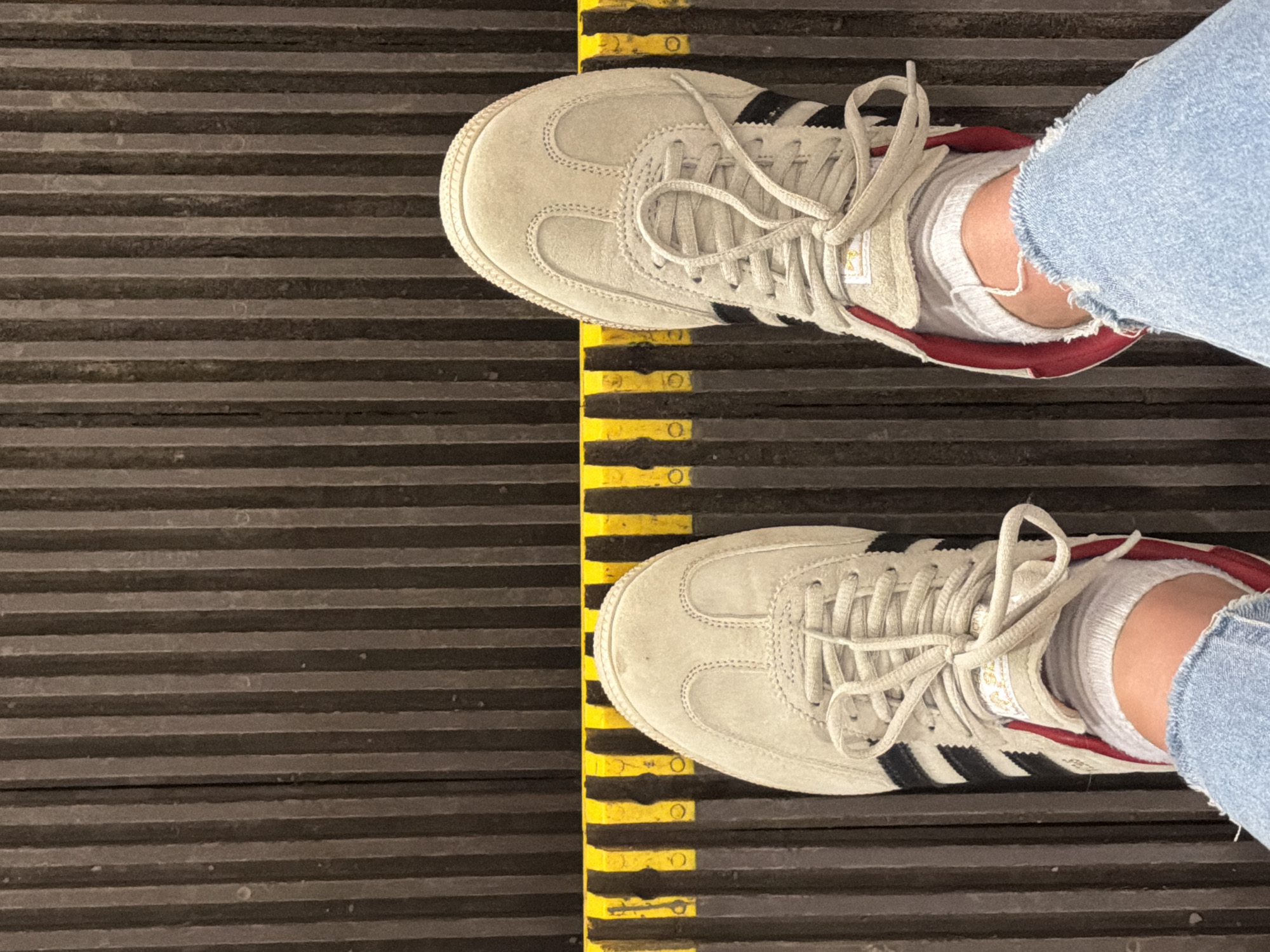 The image shows a top-down view of a person's feet wearing beige Adidas Samba sneakers with black stripes, red heel lining, and white socks while standing on an escalator. The escalator has a yellow safety stripe with round indentations. The person is wearing light blue denim jeans. The scene is potentially in Moscow, Russia, based on the common presence of these escalators in the city. The image focuses on the footwear and the escalator surface.