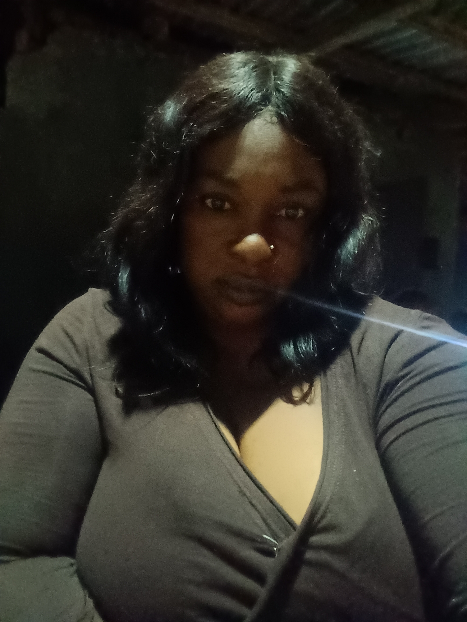A woman is seen indoors, likely in a dimly lit space in Jos, Nigeria. The woman has dark hair styled with some waves, and dark skin. A small stud earring is visible in her left nostril. She is wearing a dark brown, long-sleeved wrap top, revealing some cleavage. The background is dark and difficult to discern, suggesting an interior space with limited lighting.