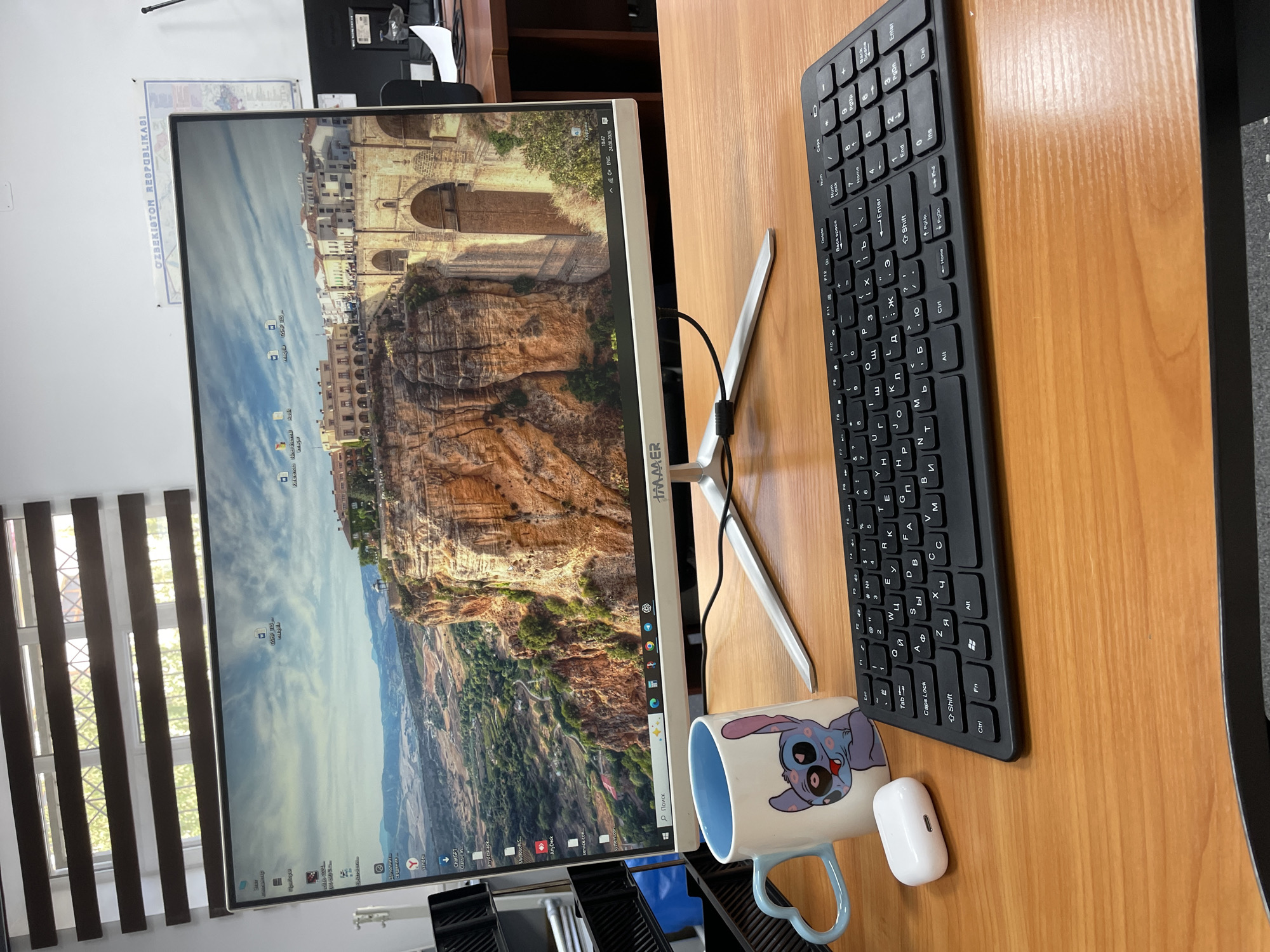 An office desk is set up with a computer monitor displaying a landscape of a city built on a cliff. The monitor is a white, flat-screen display with a silver stand. In front of the monitor, a black wireless keyboard is placed on the wooden desk. To the left of the keyboard, a blue and white mug featuring Stitch from Lilo & Stitch is positioned next to a pair of white wireless earbuds. The background shows a window with horizontal blinds and a wall with a map of Uzbekistan. The scene appears to be in an office environment in Tashkent, Uzbekistan.