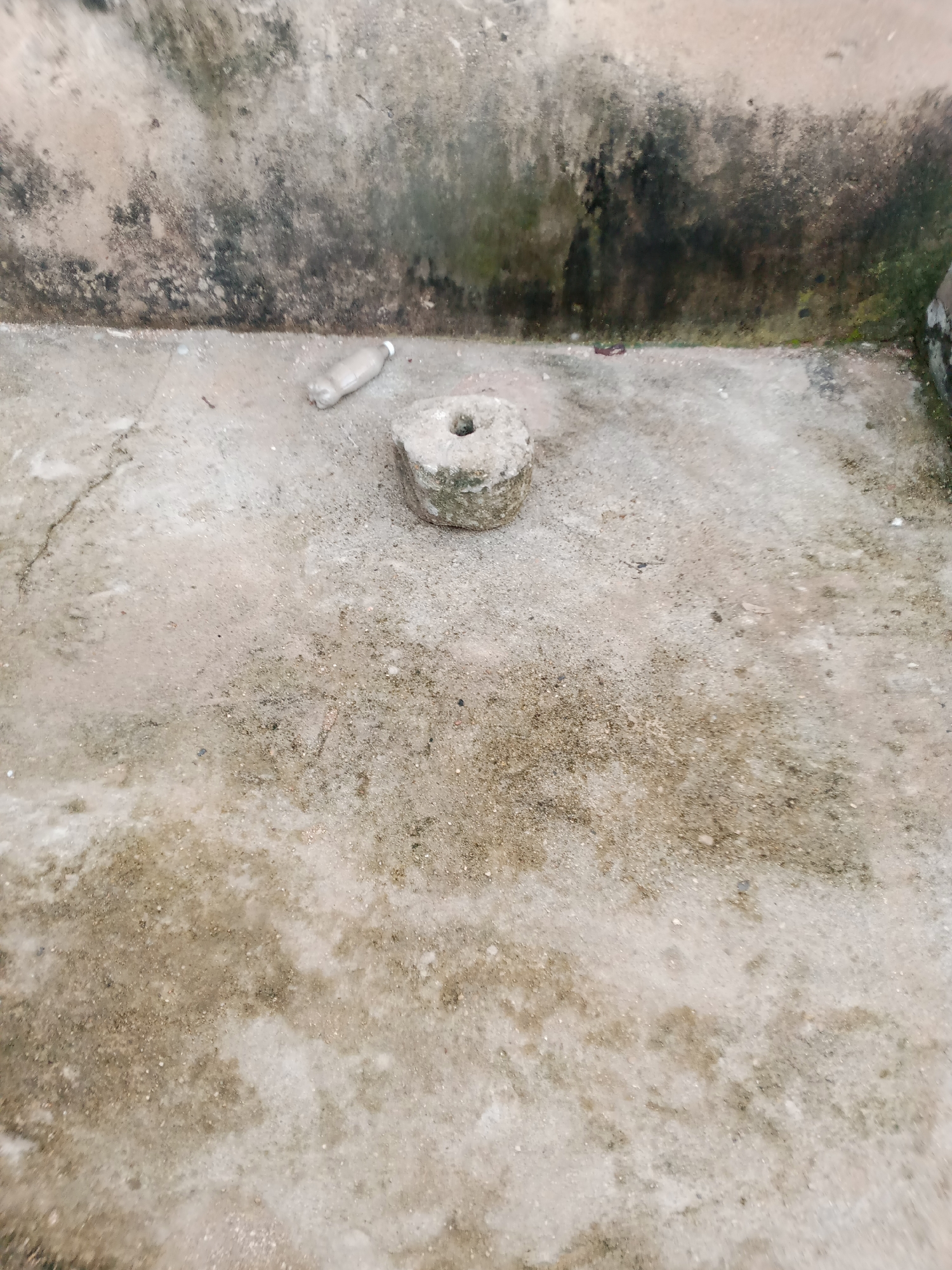 The image shows a ground-level view of a dirty, weathered concrete surface in Jauro Yinu, Nigeria. A small, circular stone with a hole in the center is prominently positioned in the foreground. To the left of the stone, a used plastic bottle lies on the ground. The background features a rough, stained wall with patches of dark mildew and green algae. The overall scene suggests an outdoor, possibly neglected area within the city.