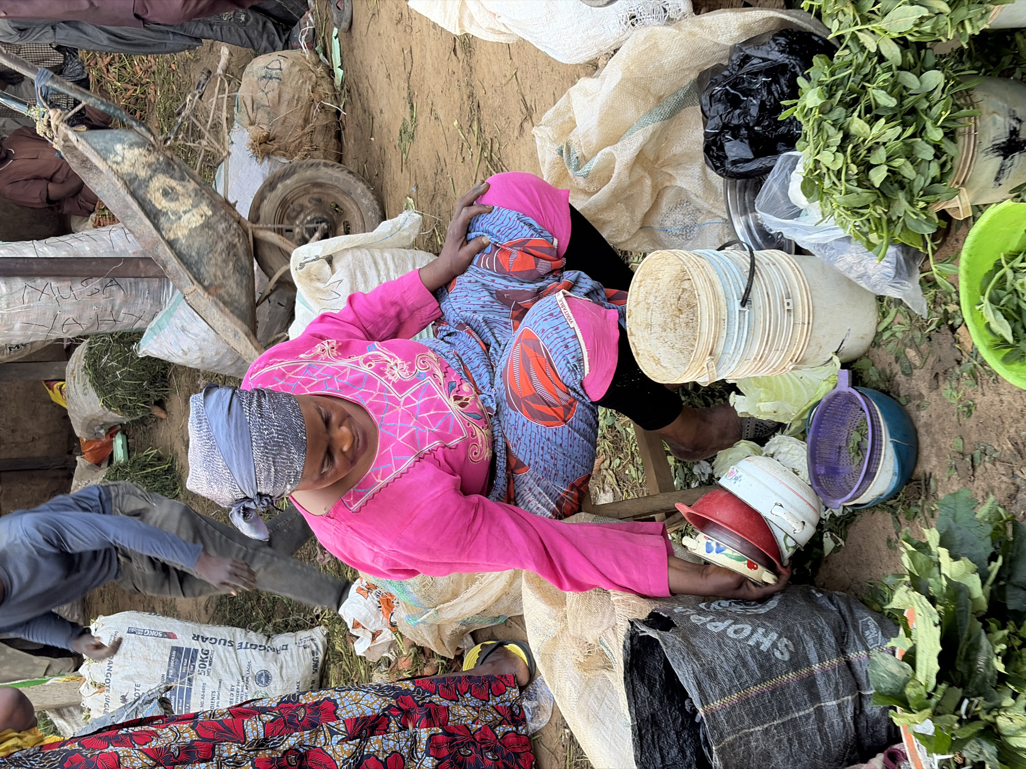 In Gura Top, Nigeria, a woman wearing a bright pink top and a blue and red patterned skirt is seated on a stool. She is reaching for a stack of bowls, including a red one and a white one with floral designs. Surrounding her are various items, including a large black bag with "SHOPPING" printed on it, a pile of green leafy vegetables, and several white buckets. A man in a blue long-sleeved shirt and dark pants is visible in the background, standing near a large white bag of sugar. A wheelbarrow loaded with produce is also present in the scene, suggesting a market or trading environment. The ground is dusty and scattered with debris.