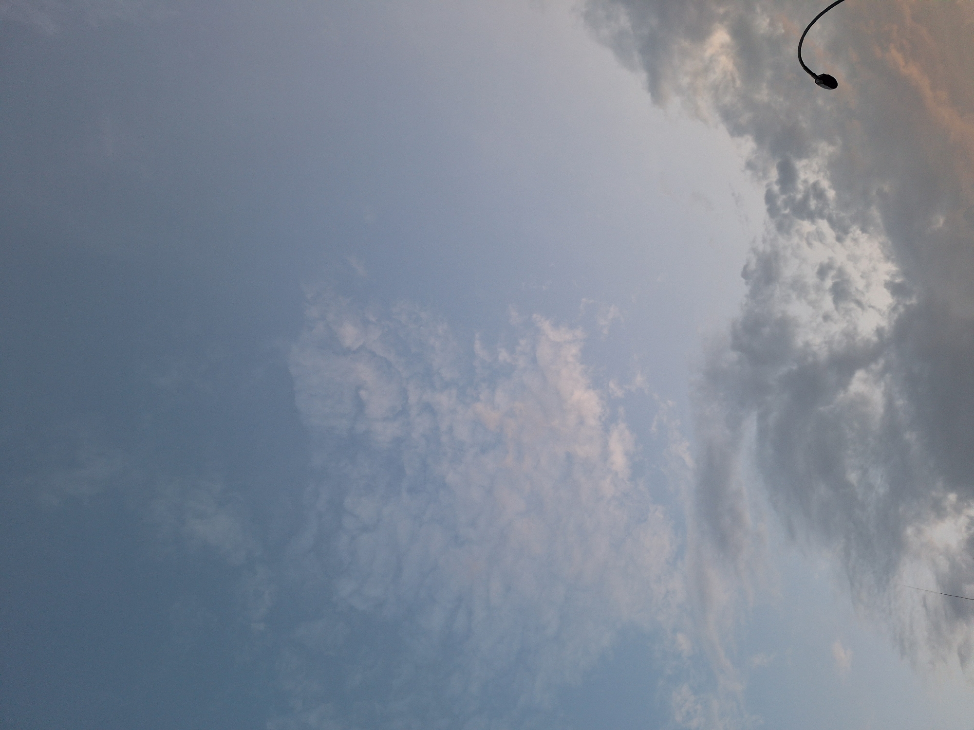 The image shows a cloudy sky with a streetlamp on the right side. The sky is a soft blue and grey, with hints of pink and orange from what appears to be a sunset or sunrise. The clouds are varied, with some thin and wispy, while others are darker and more substantial. The streetlamp is black and curves towards the left, with its light fixture visible at the end. The image appears to be taken from a low angle, looking up at the sky. There are no people or other identifiable objects in the scene. There is no indication of the location within the city of Prague, Czechia.