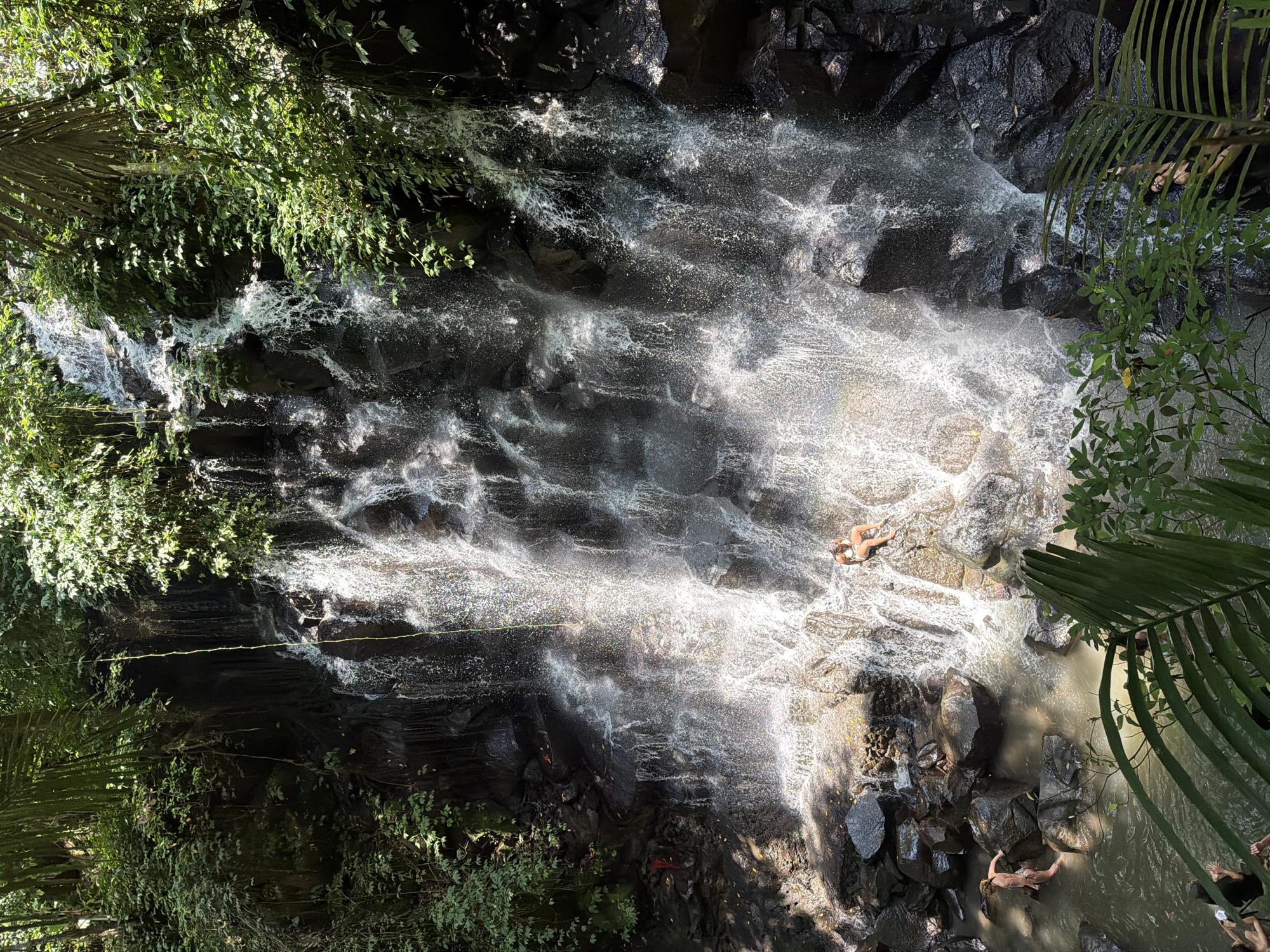 The image shows a waterfall in Beng, Indonesia. The waterfall cascades over dark rocks, creating white foam and spray. The water collects in a pool at the bottom where people are bathing. Green foliage surrounds the waterfall. A safety rope stretches across the rocks next to the falls.