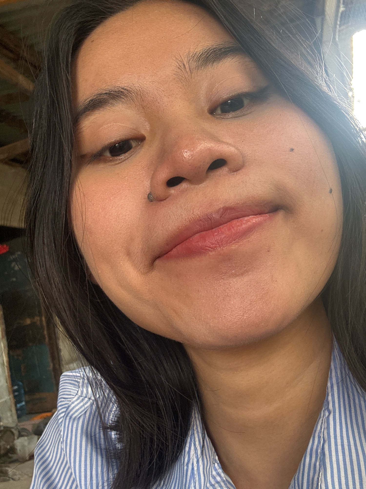 The image is a close-up selfie of a young Asian woman. She has dark hair, brown eyes, and is wearing a blue and white striped shirt. Her face is angled slightly upwards and to the side, with her lips slightly parted. There is a small mole on her left cheek and a piercing on her left nostril. The background appears to be outdoors, with some blurred structures and foliage visible. The lighting suggests it is daytime. No specific location in East Jakarta, Indonesia can be determined from the image.