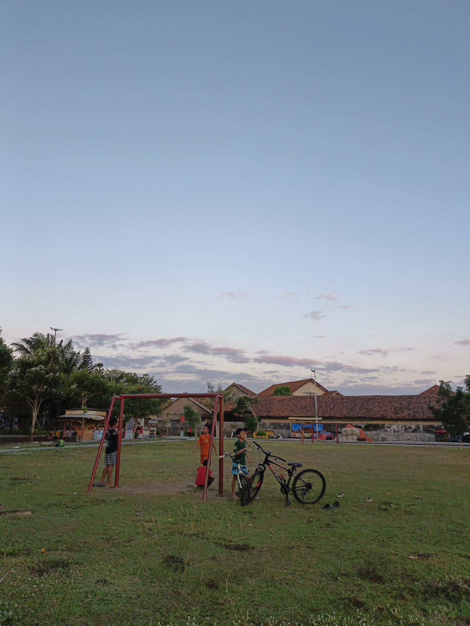 In Klaten, Indonesia, a park scene unfolds under a pale blue sky with wispy clouds. The foreground is a grassy field with scattered patches of wildflowers. A red metal soccer goal stands center-left. Three boys are near the goal; one in a black shirt and checkered shorts stands to the left, another in an orange shirt is in the middle, and a third in a green shirt is to the right. A bicycle with a black frame and red accents leans to the right of the green-shirted boy. Two pairs of sandals are on the ground near the bicycle. In the background, a cluster of trees frames a row of buildings with terracotta tile roofs, suggesting a residential or community area. Two small food stalls are visible behind the soccer goal. The overall atmosphere is casual and recreational, with children engaging in outdoor activities.