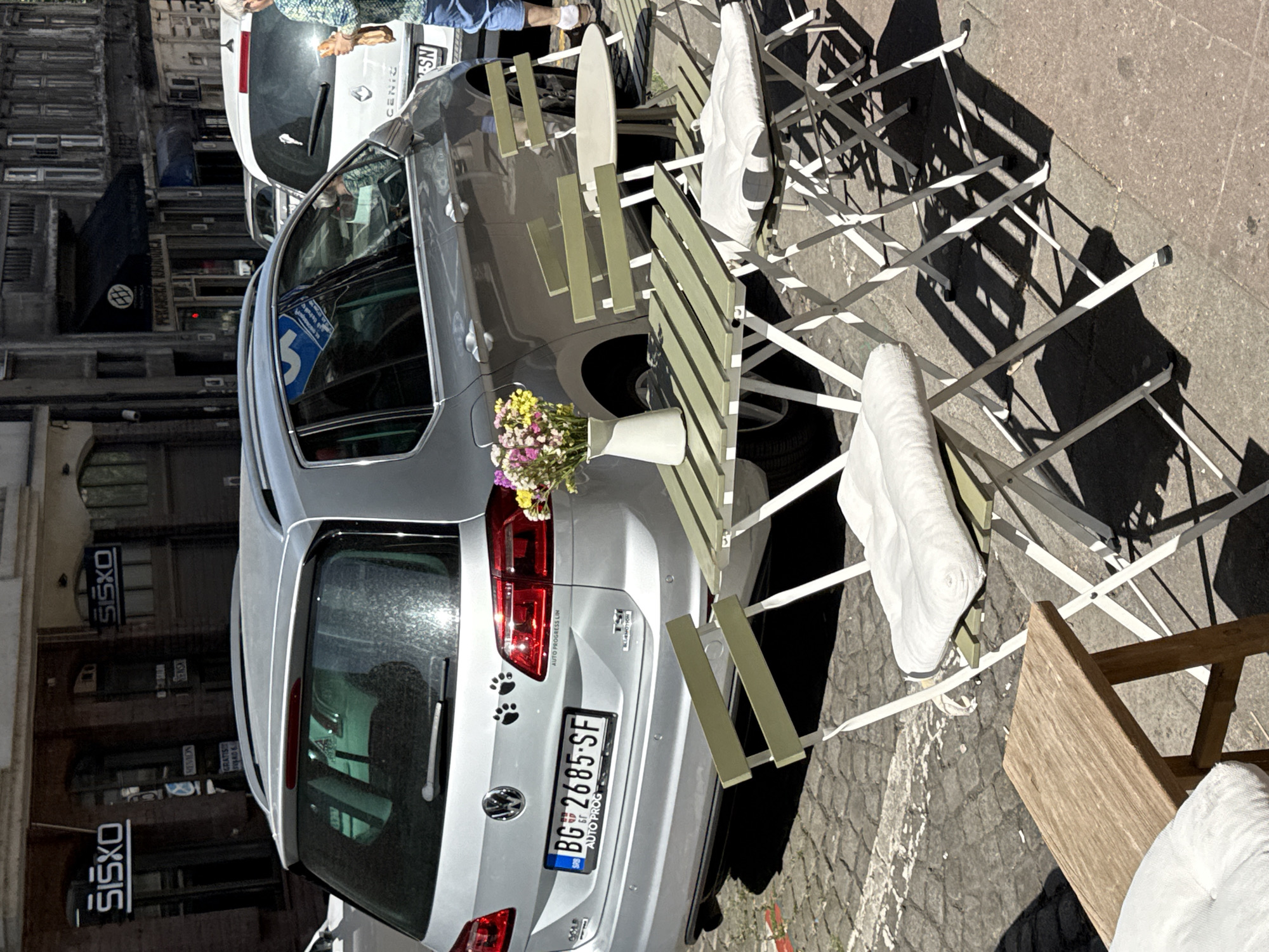 The image captures a street scene, likely in Belgrade, Serbia. A light grey Volkswagen Golf is parked alongside outdoor cafe tables and chairs, some with white cushions. A white Renault Scenic is partially visible behind the Golf. A vase with flowers sits on the Golf's rear bumper. Paw print stickers are affixed to the car's rear window. The car's license plate reads "BG 2685-SF", indicating Serbian registration. Shop signs for "SISXO" and "PEKARSKA KLAVNICA" (bakery and slaughterhouse) are visible on nearby buildings. Pedestrians are partially visible near the Renault Scenic. The scene is bathed in sunlight.