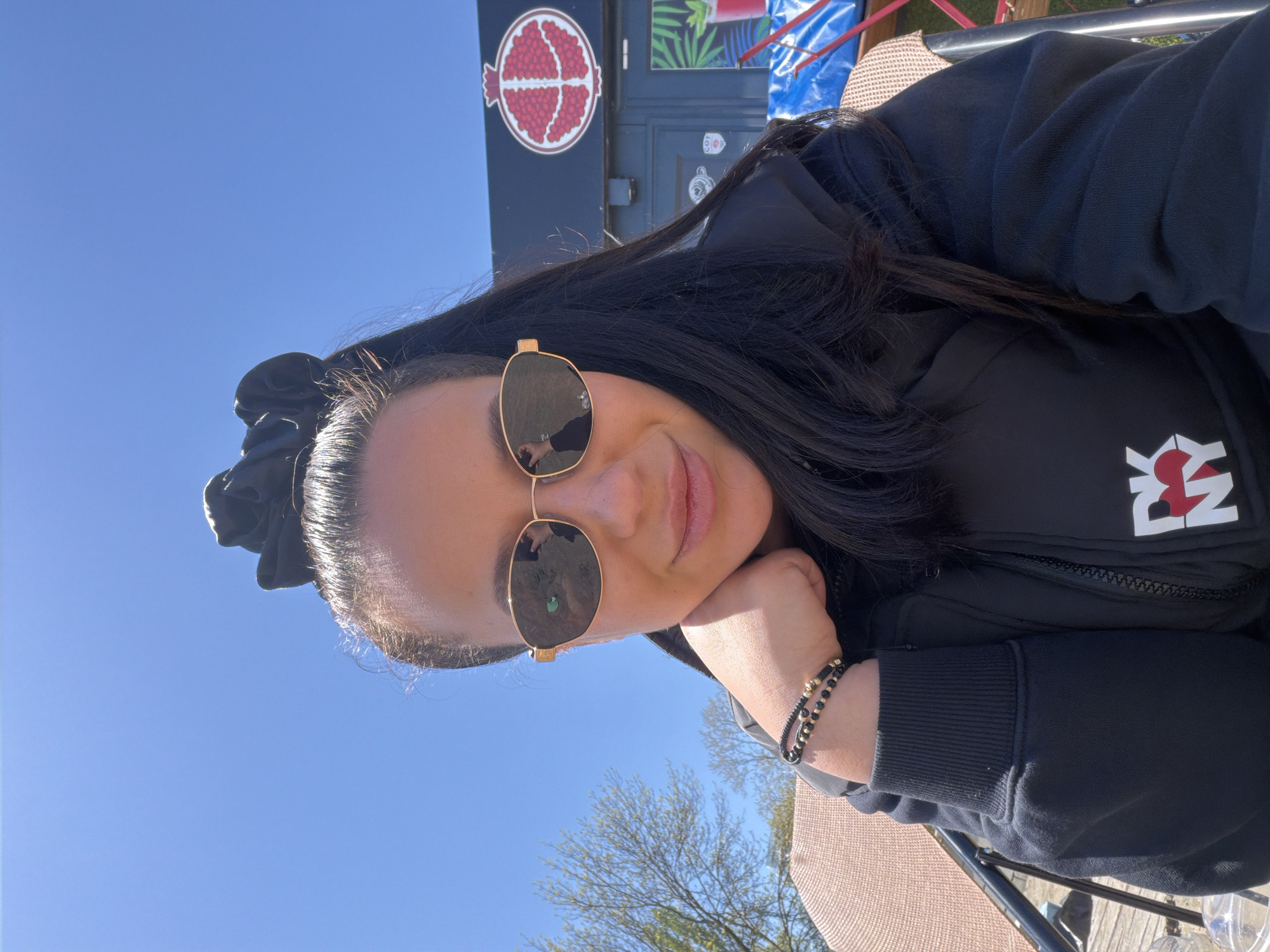 A woman is seated outdoors on a chair or bench, with a clear blue sky partially visible above. She is wearing a black jacket with a white logo on the left chest and gold-framed sunglasses. A black hair accessory is visible in her hair. She rests her cheek on her hand, adorned with two beaded bracelets. In the background, there is a food truck or trailer with a pomegranate logo and tropical plant artwork.