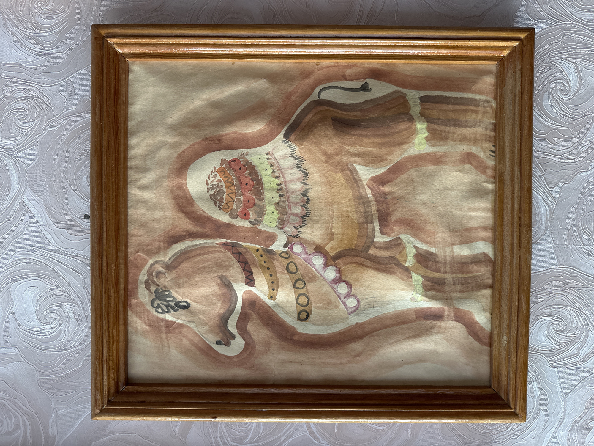 A framed watercolor painting is centered in the image against a textured white background. The painting depicts an abstract brown figure with ornate decorations. The figure has a decorated headpiece with red, orange, yellow, and green details. Markings and patterns are visible on the body. The painting is contained within a wooden frame with a gold finish. The scene is located in the city of Sayansk, Russia.
